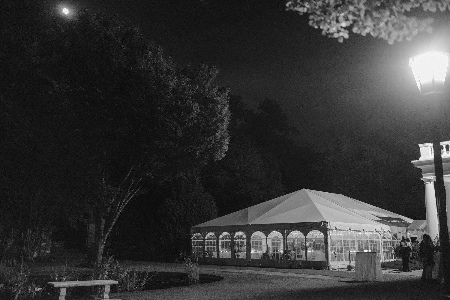 Black and white photo of an illuminated wedding tent at night with a glowing lamp post.