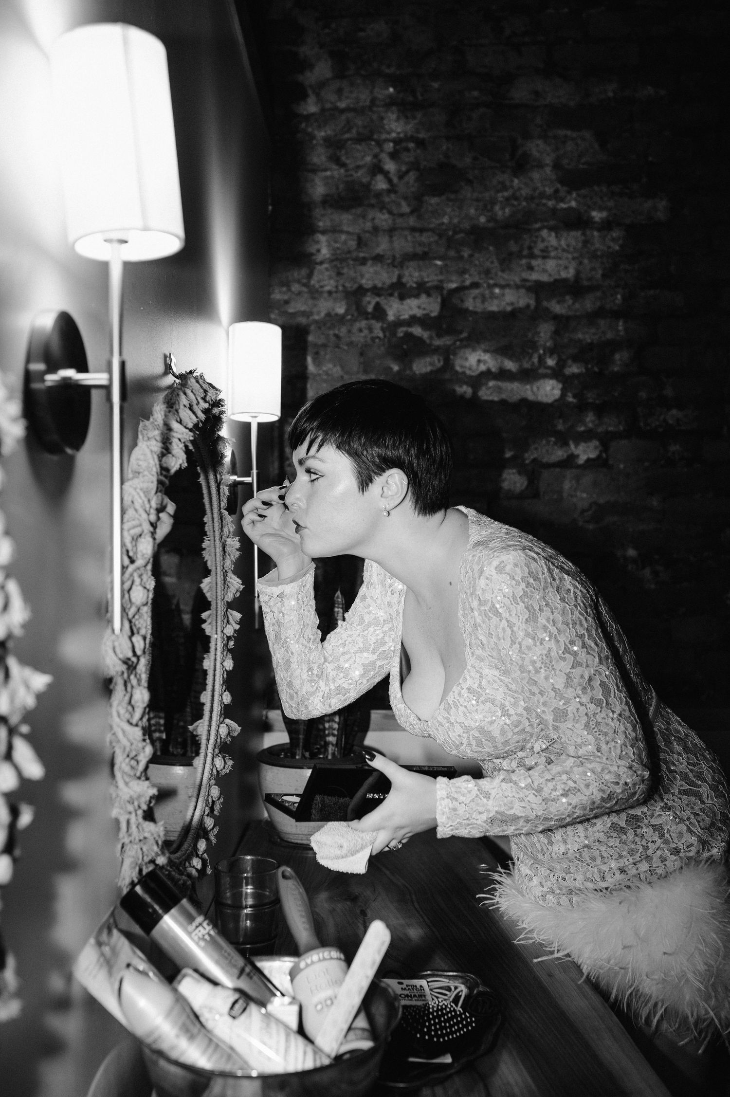 A person applies makeup in front of a mirror with wall sconces in a brick-walled room.