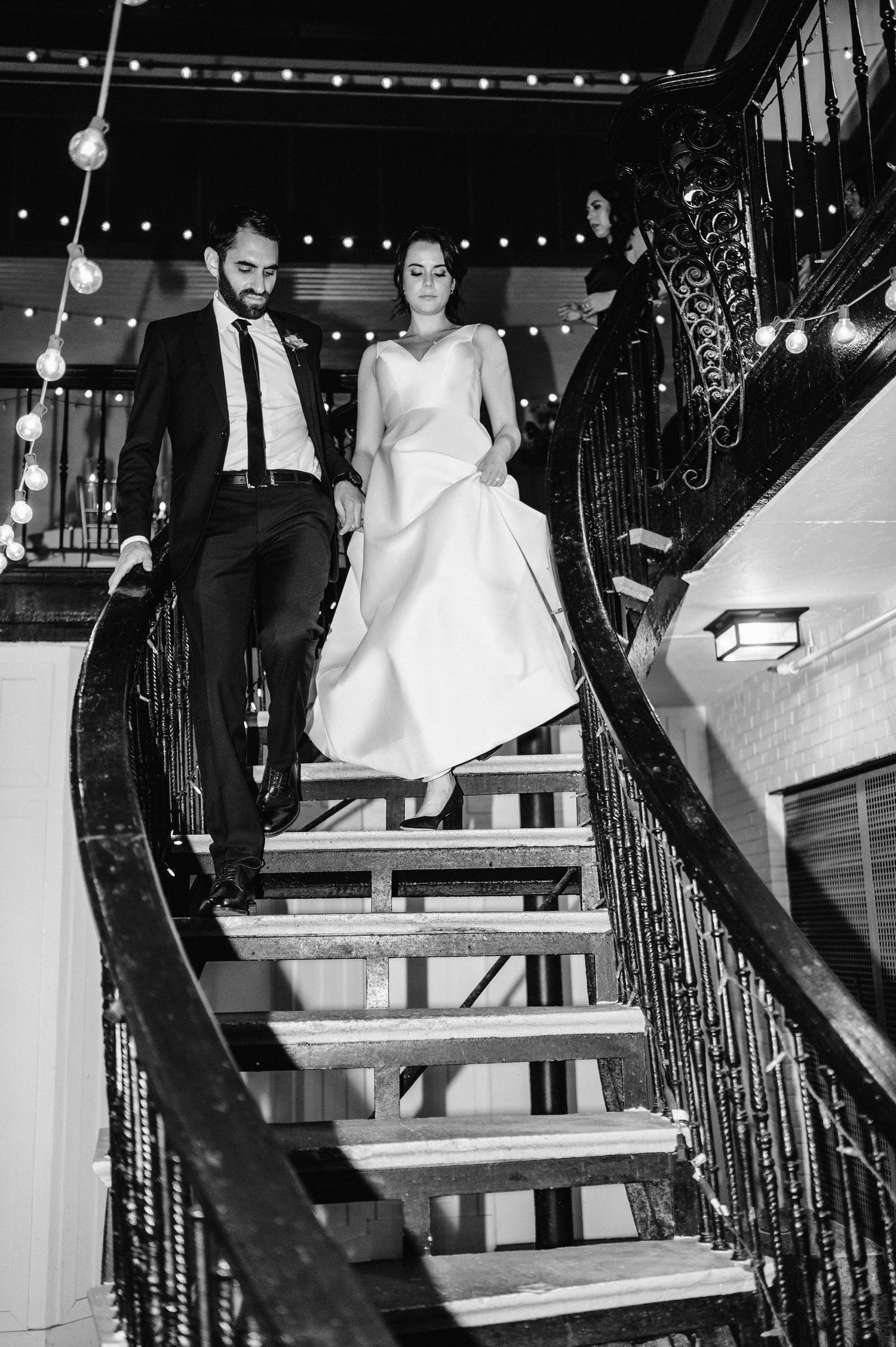 Couple descending ornate iron staircase under string lights at wedding venue.