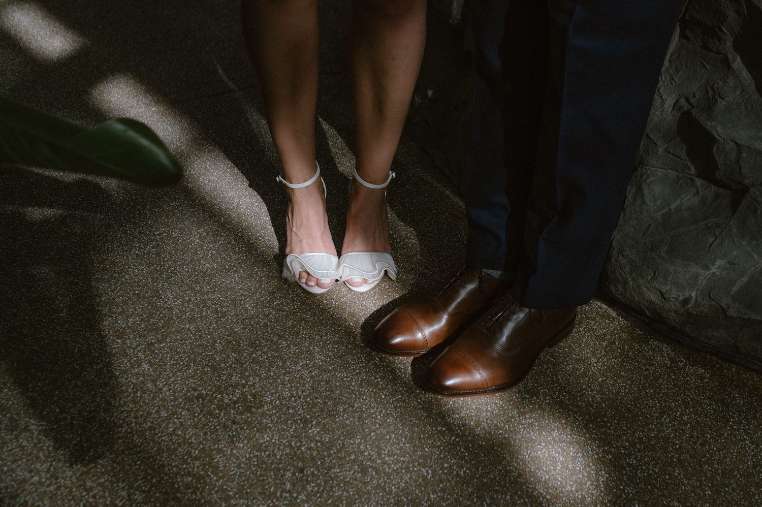 Close-up of white bridal shoes and brown dress shoes standing together on dark flooring.