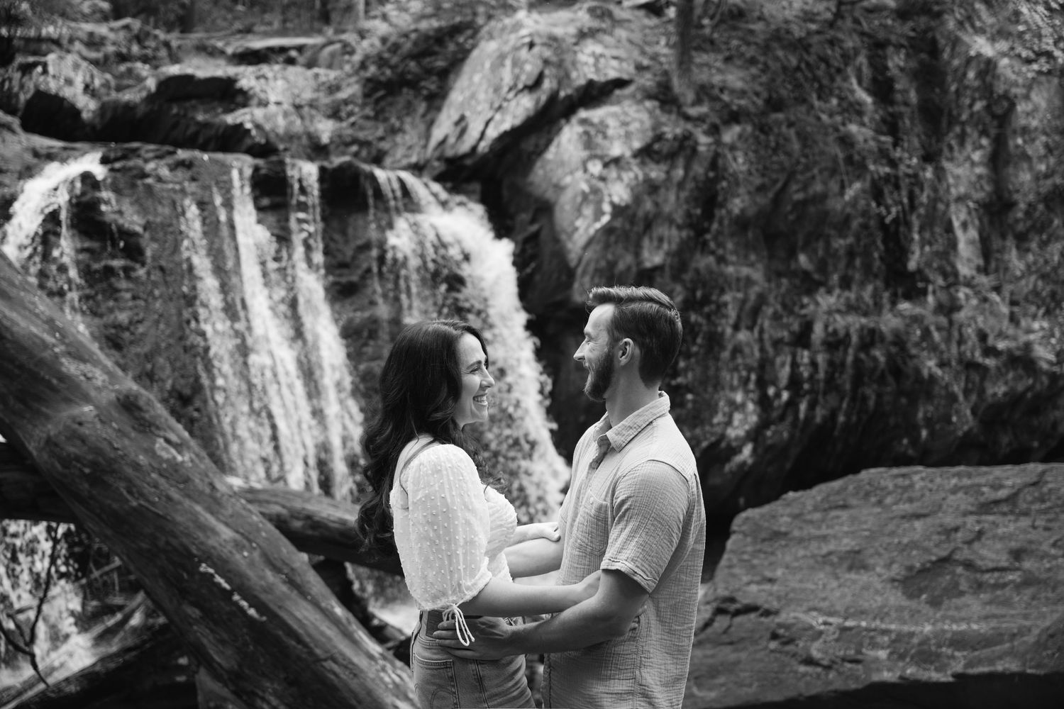 Black and white photo of a couple holding hands in front of a waterfall surrounded by rocks and logs.