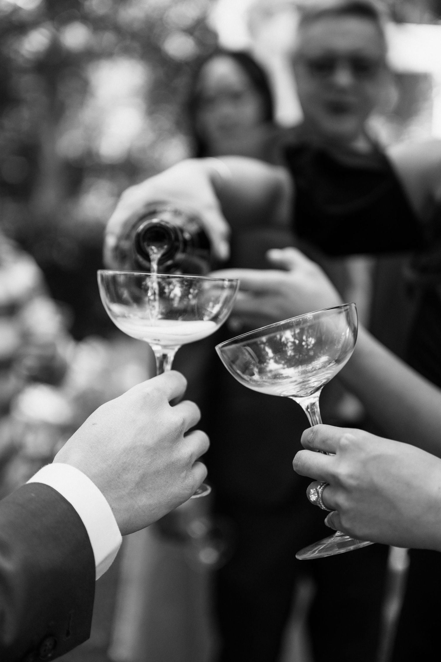 Black and white photo of hands holding champagne coupe glasses while being poured at an elegant celebration event.