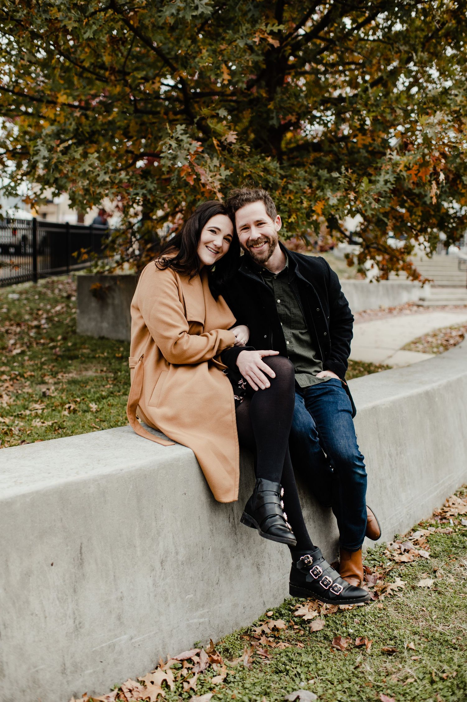 A smiling couple sits together on a concrete wall outdoors in autumn, woman in camel coat, man in dark jacket.