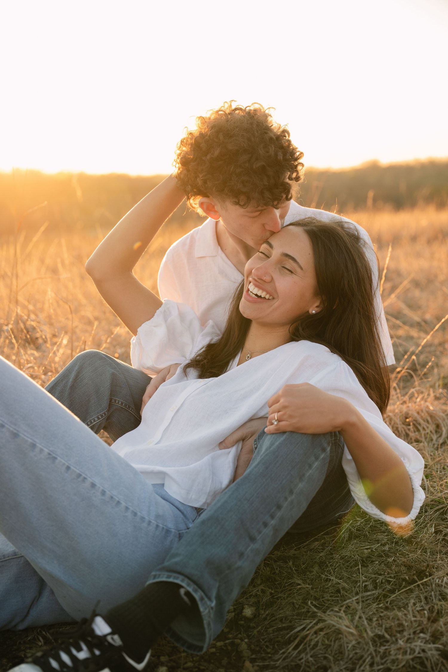 Couple sharing a romantic sunset moment in a golden field, woman laughing joyfully as man kisses her head.
