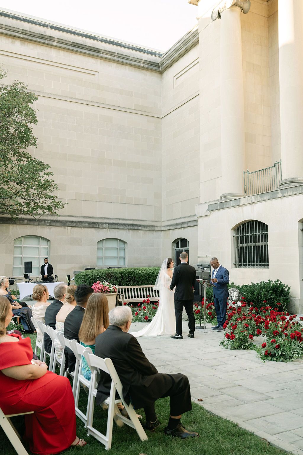 A wedding ceremony takes place on an elegant stone patio with white columns and red flowers in the background.