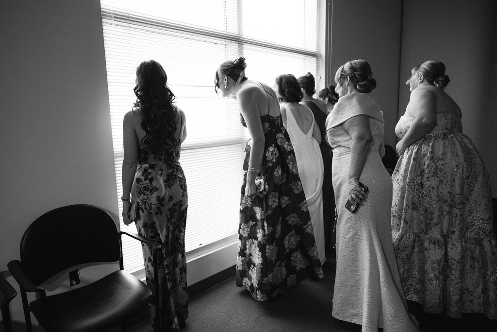 Black and white artistic photograph showing silhouettes of wedding dresses hanging in a row by a window.