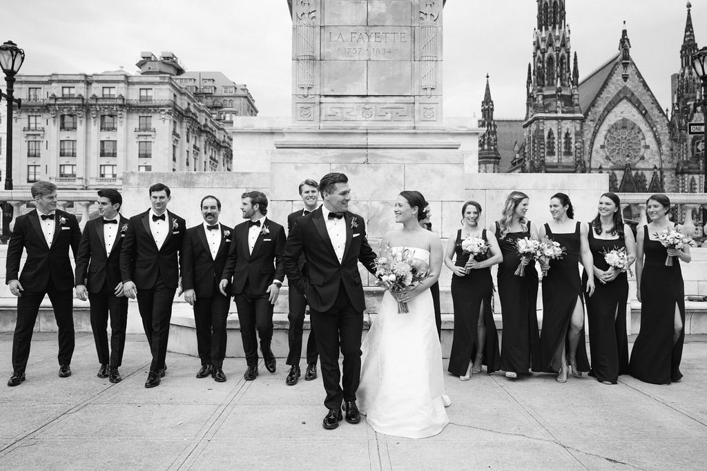 Wedding party in formal attire poses in black and white against architectural backdrop with church spires.