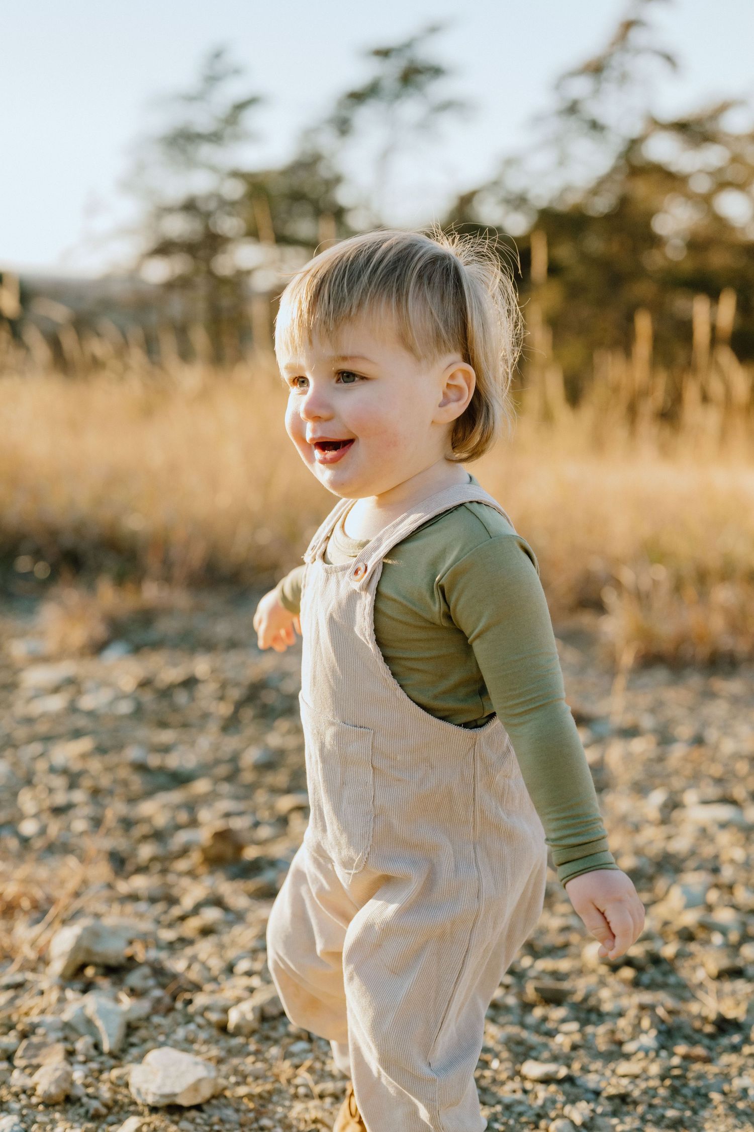 Toddler in overalls and green shirt smiles while playing outdoors.