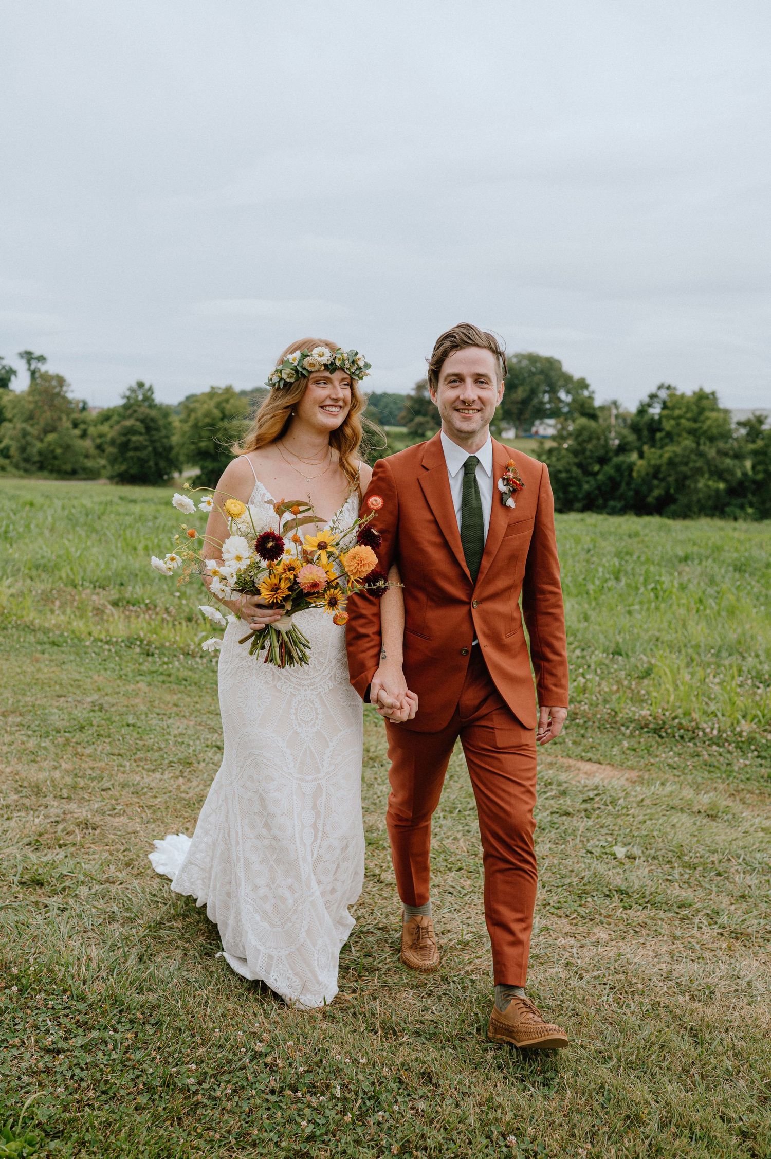 Wedding portrait in field with bride in lace dress and groom in rust-colored suit.