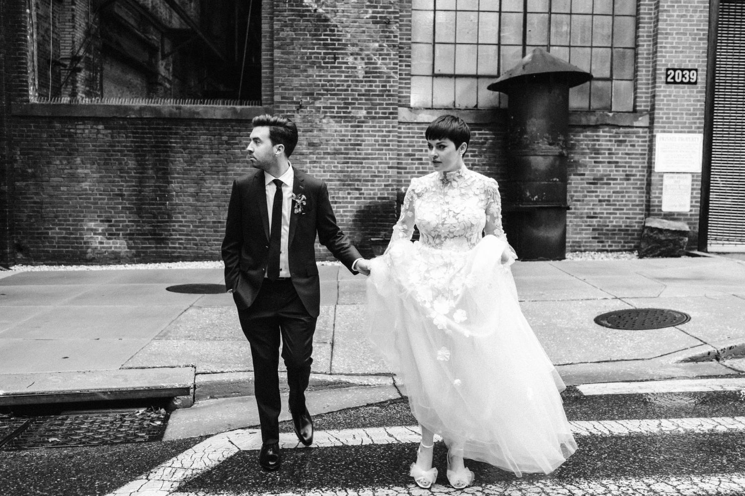 A couple walks together on a cobblestone street outside an industrial brick building on their wedding day.