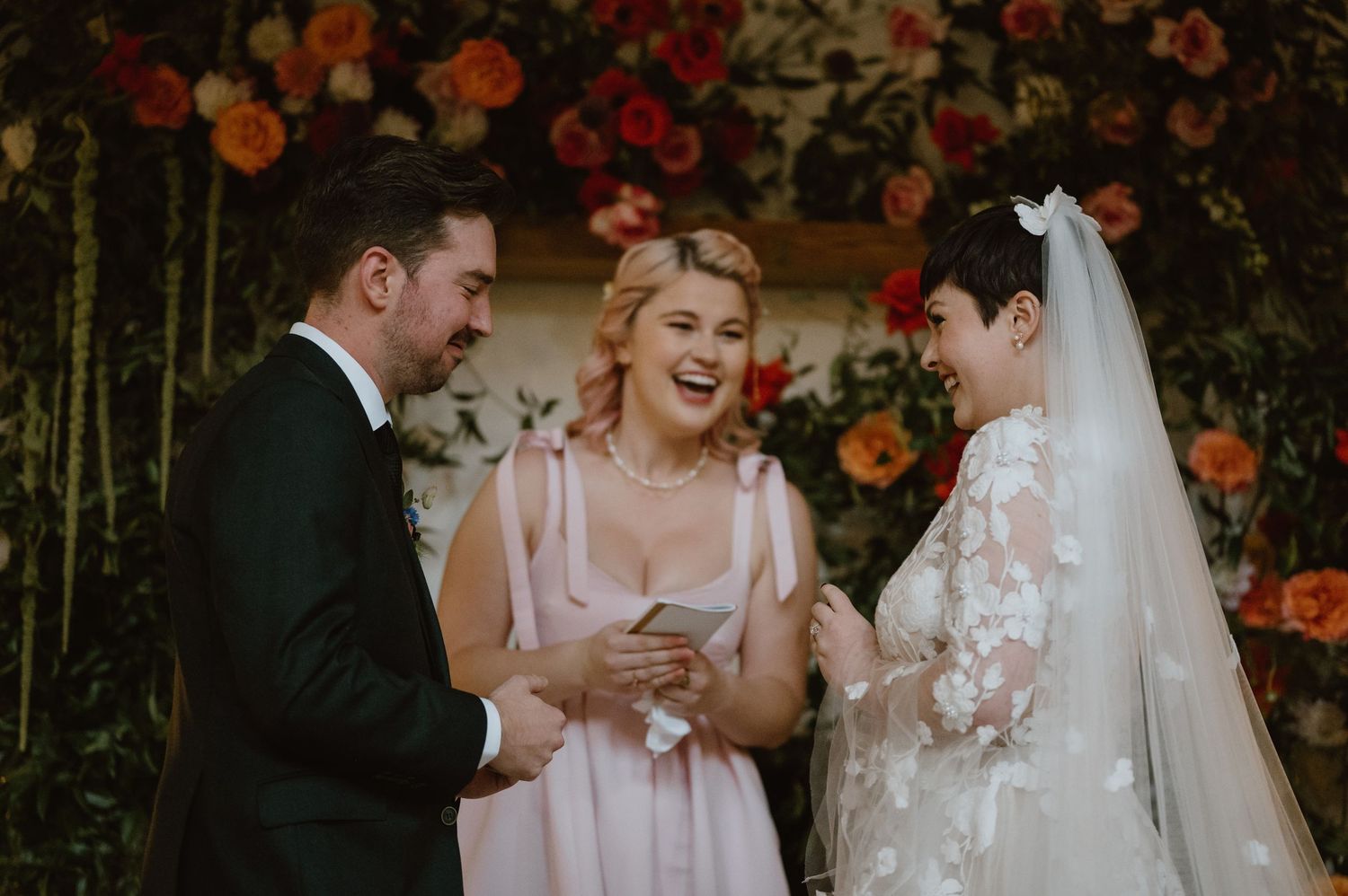 A wedding ceremony moment shared under a floral arch with orange and red flowers.