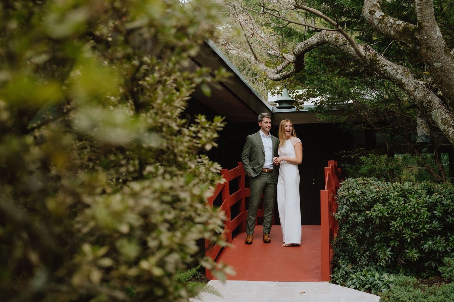 Couple standing on red porch of rustic cabin surrounded by lush greenery.