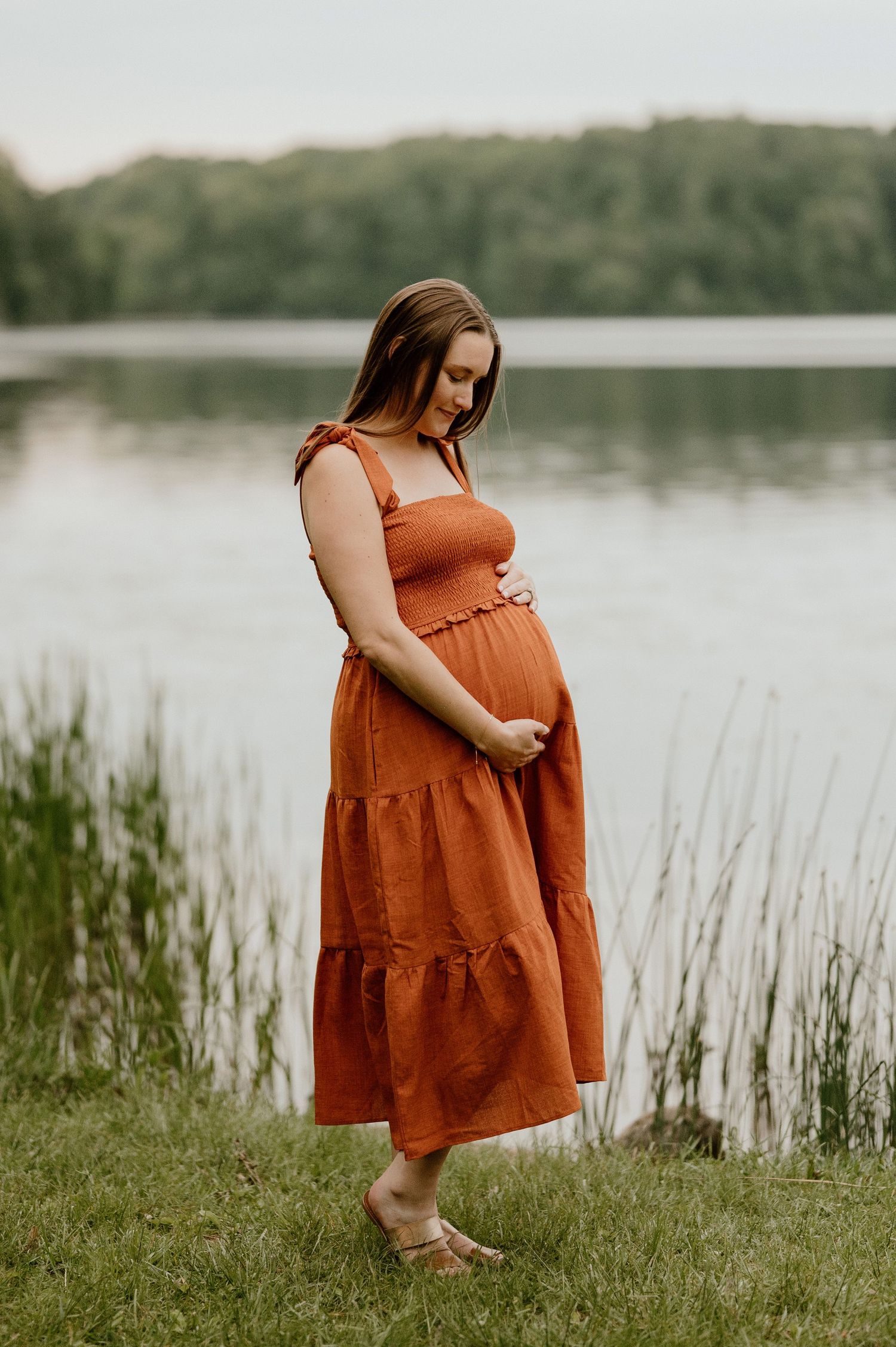 An expectant mother in a tiered orange dress stands thoughtfully by a serene lake.