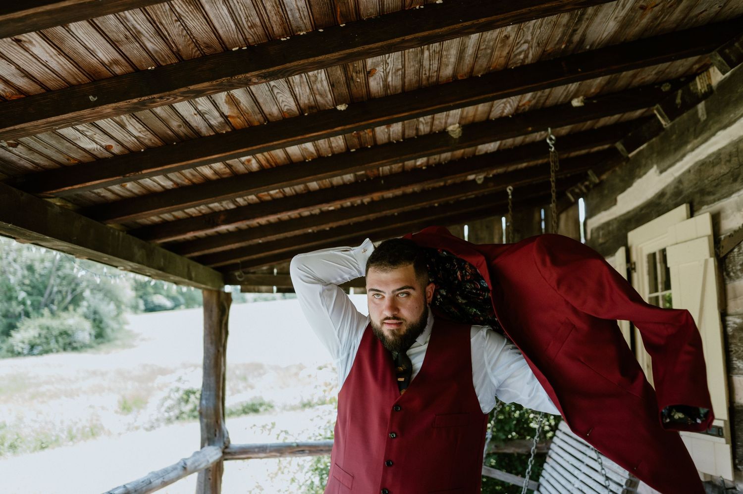 Two people in burgundy formal wear playfully pose under a rustic wooden porch.