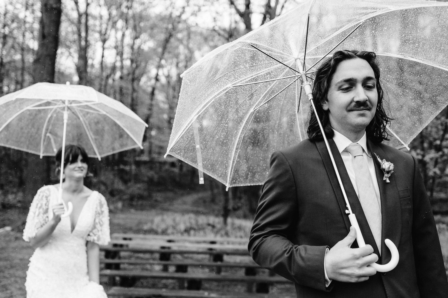 Wedding couple with clear umbrellas stand apart during outdoor forest ceremony.