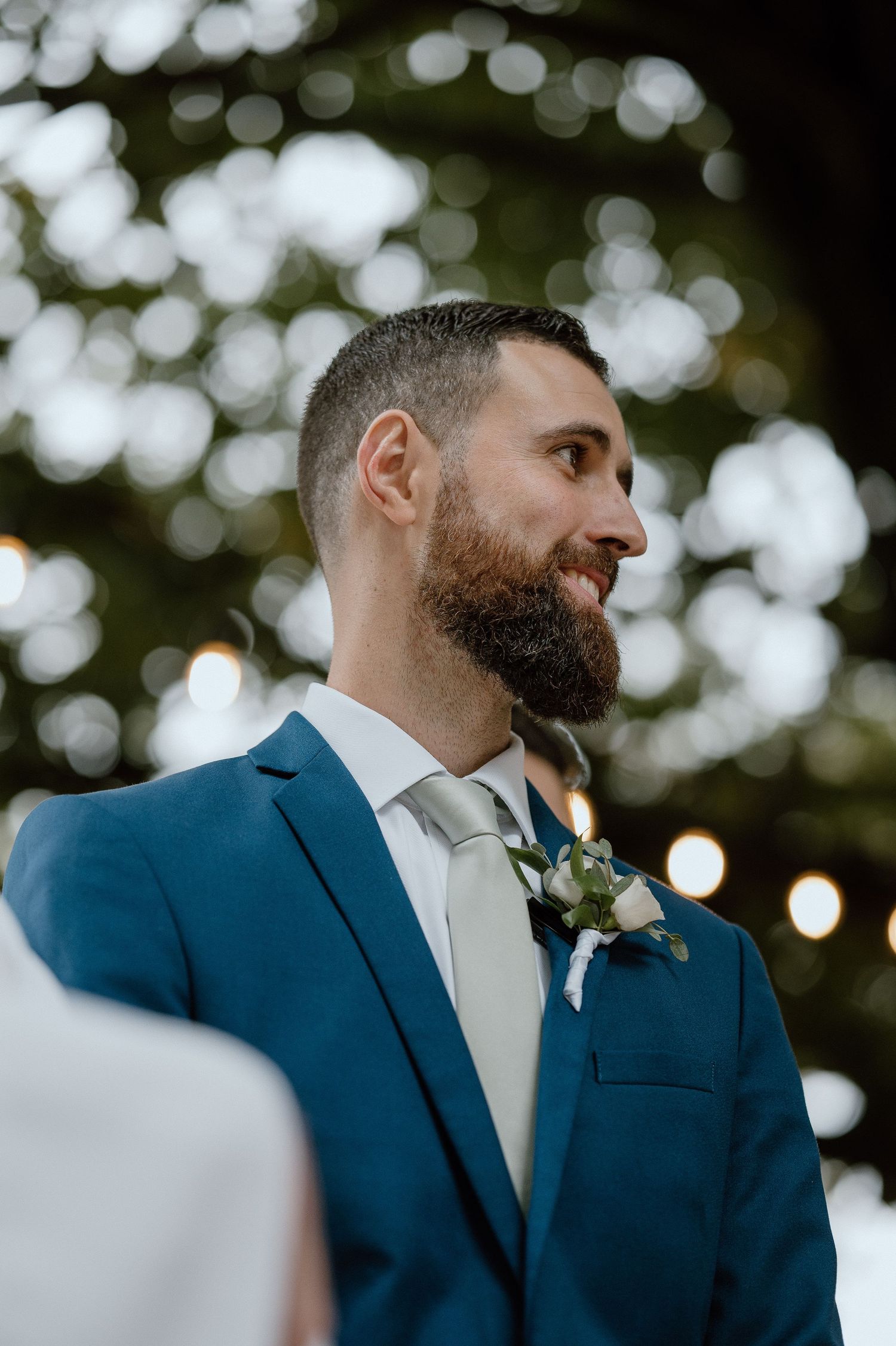A groom in a navy blue suit stands in an outdoor setting with bokeh lights in the background.