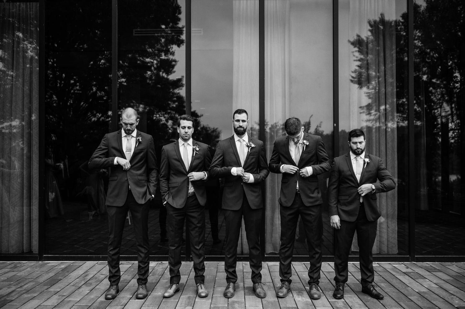 Groomsmen in formal attire pose together against modern glass building exterior.