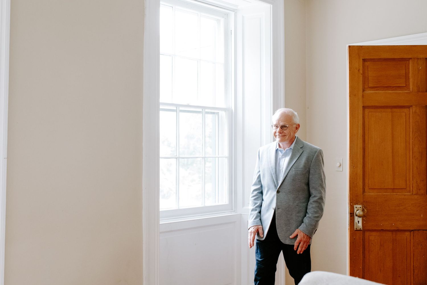 A person in a light gray suit jacket stands near a bright window in an elegant room.