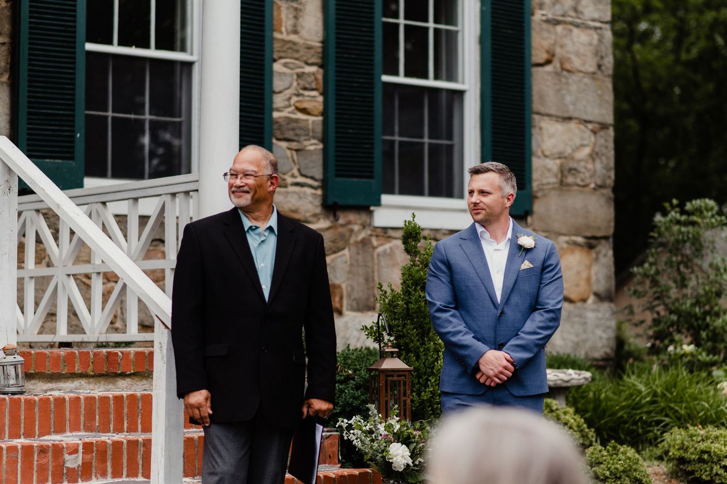 Two people standing on brick steps of historic stone house during outdoor ceremony.