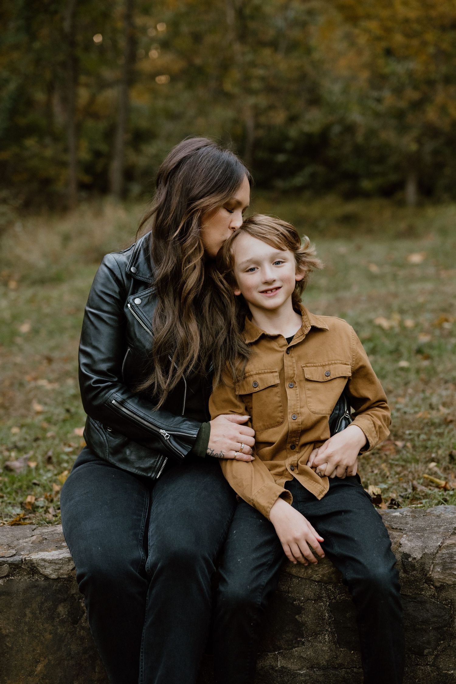 Two people share a tender moment while sitting on a stone wall surrounded by autumn foliage.