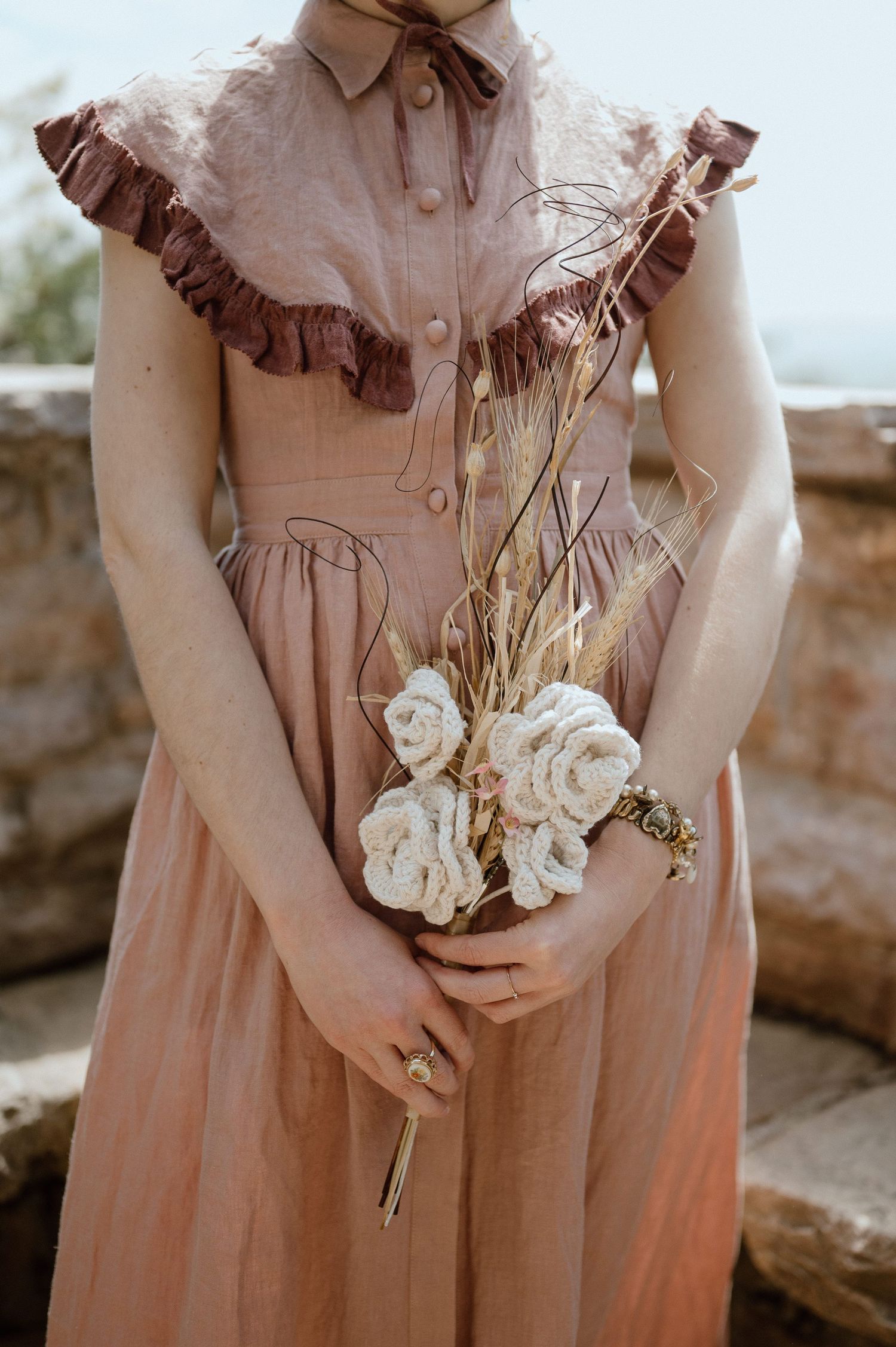 A feminine dusty pink vintage-style dress with ruffled details holds a bouquet of white roses against a stone wall.
