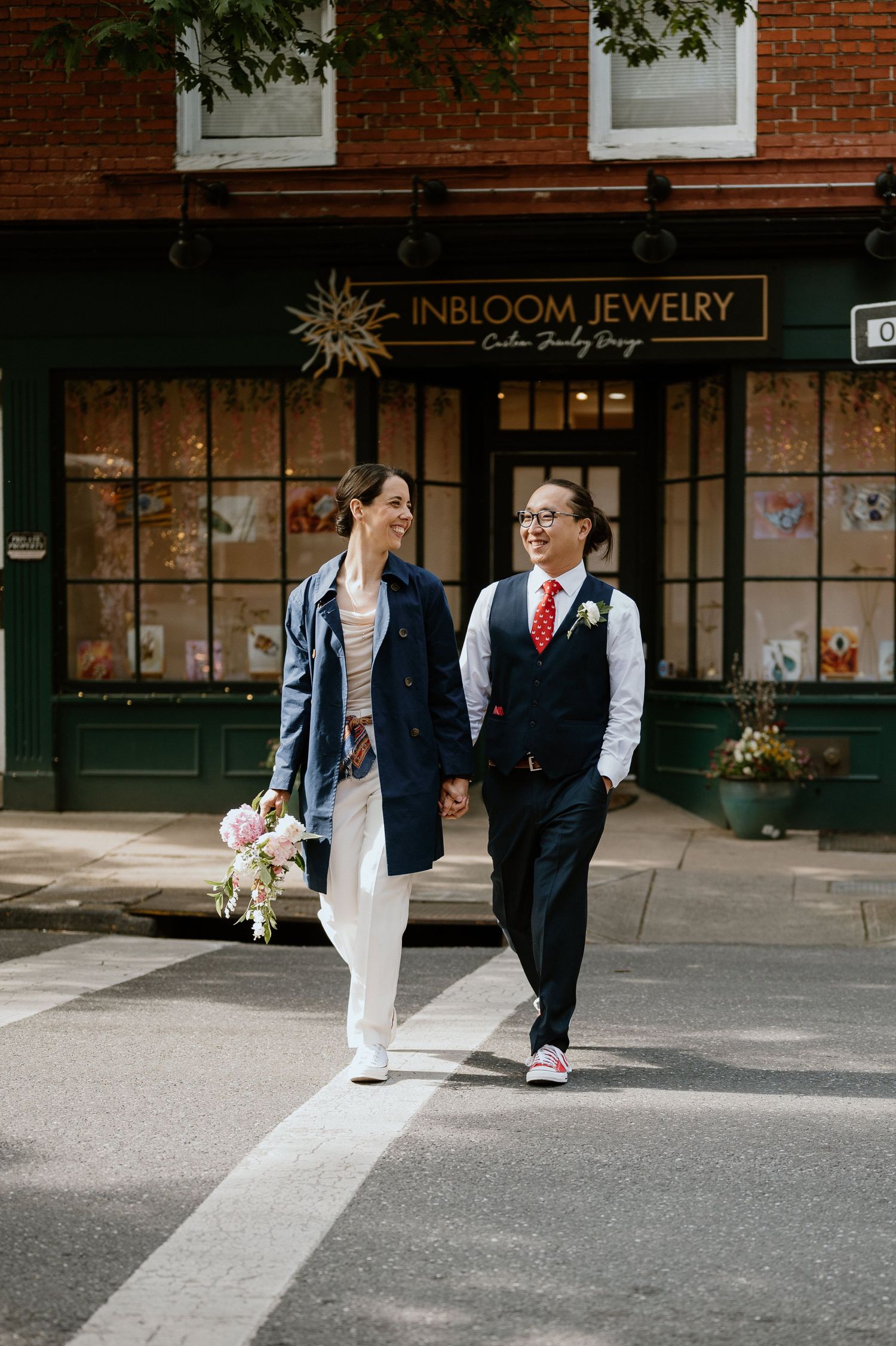 A couple walks hand in hand past InBloom Jewelry store in formal attire against a brick building backdrop.