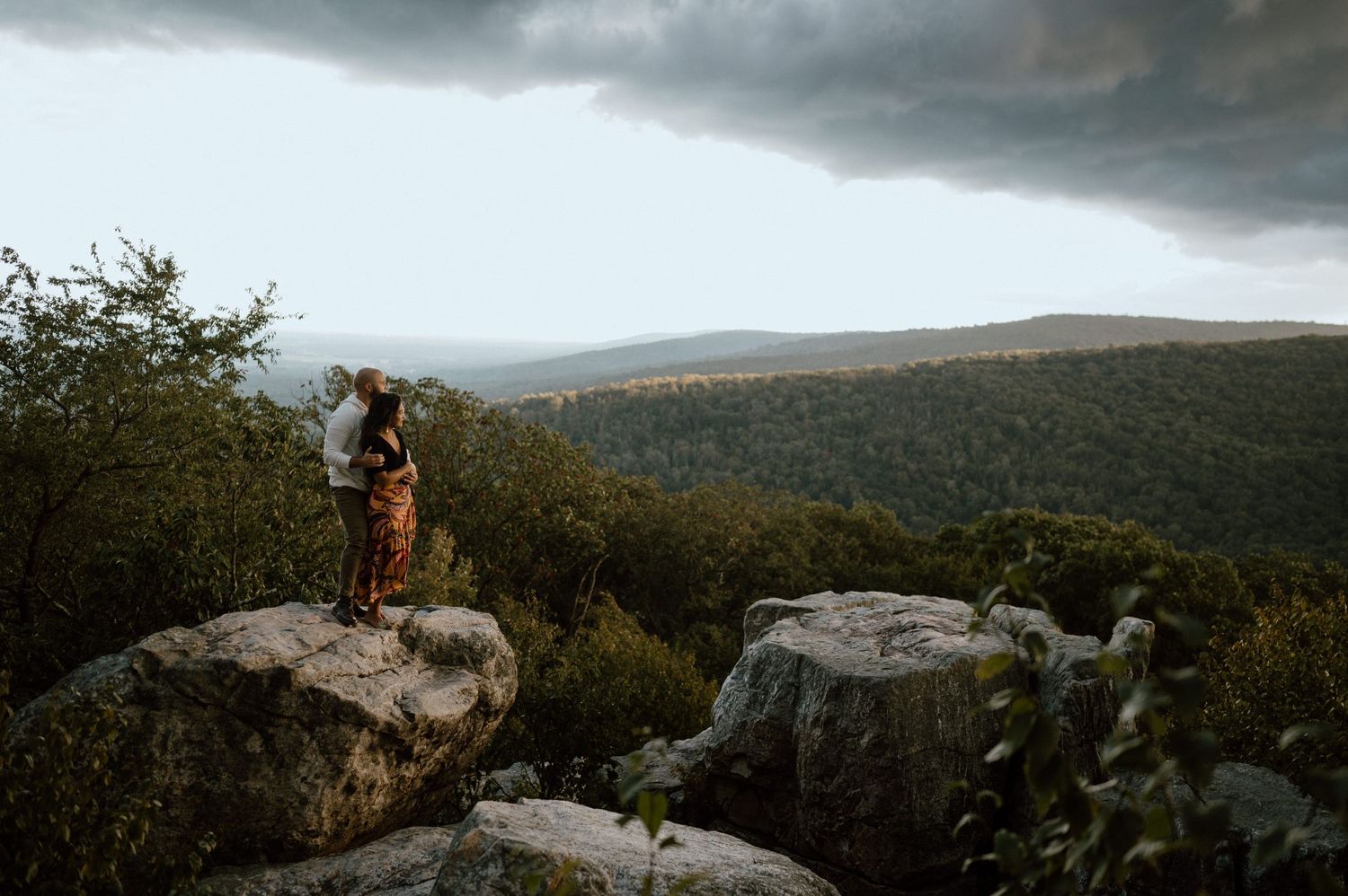 A couple stands together on dramatic mountain rocks with stormy skies and forest views behind them.