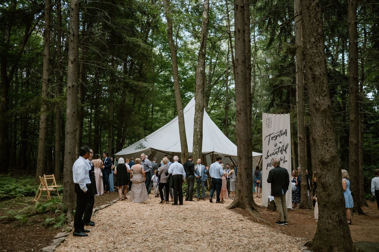 Wedding guests gather under a white tent surrounded by towering forest trees.