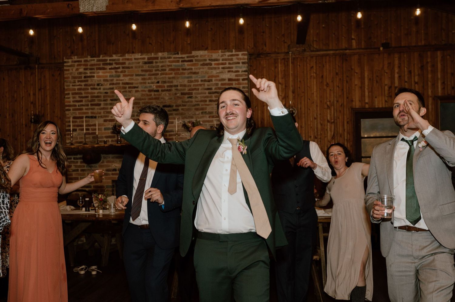 Wedding guest in green suit dancing enthusiastically at reception with others in background.