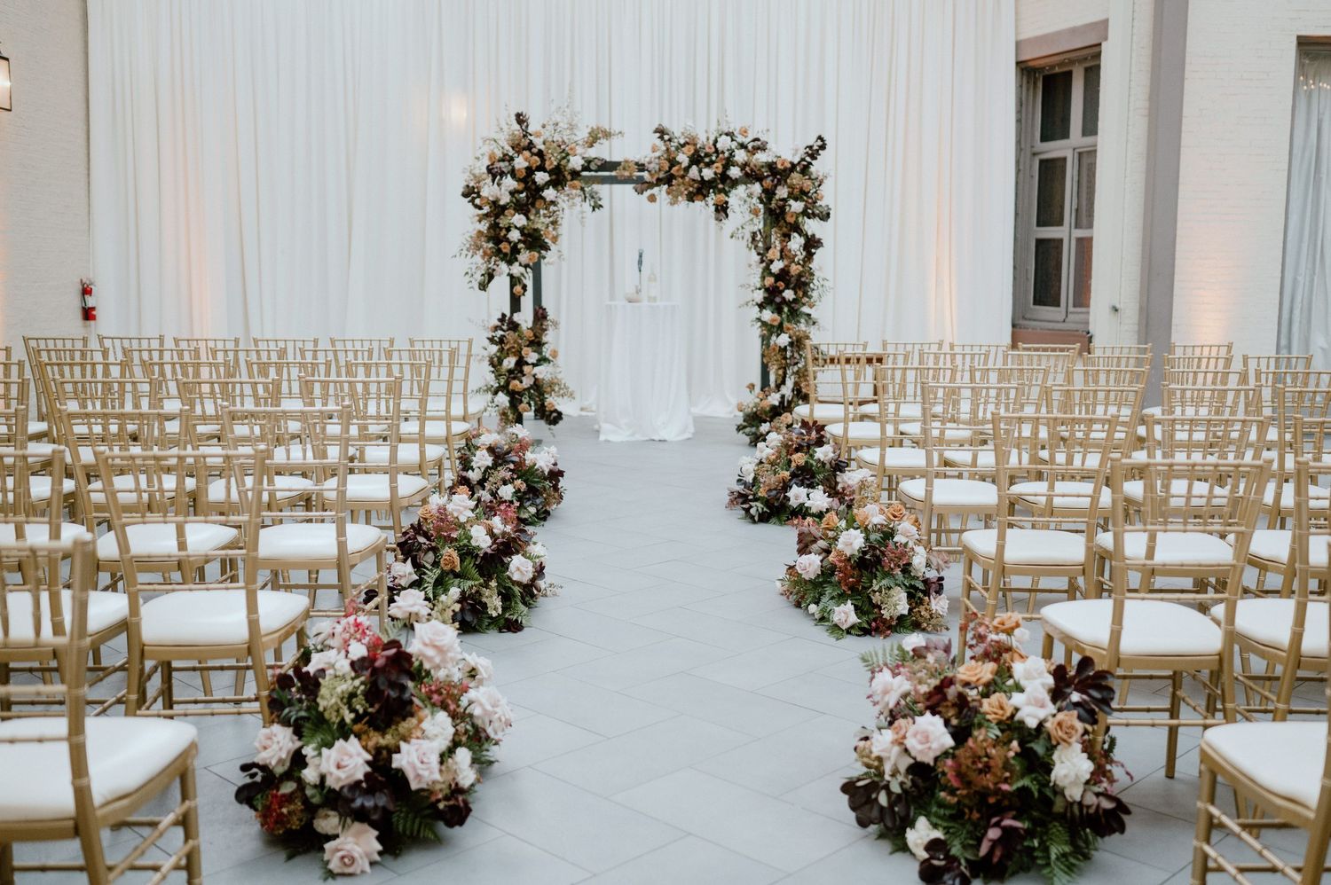 Wedding ceremony setup with floral arch and gold chiavari chairs in white room.