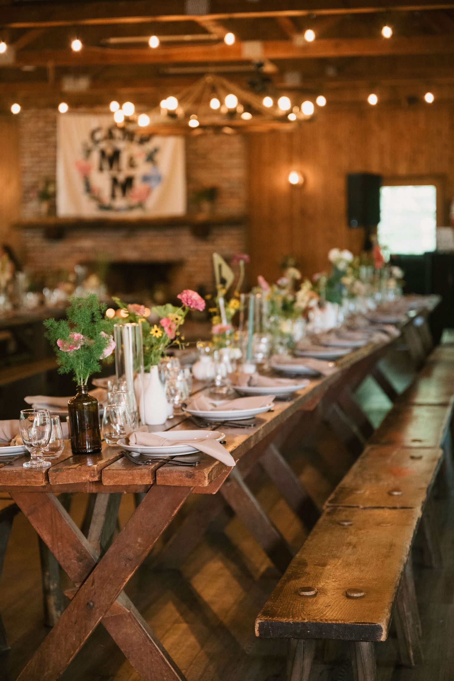 Long wooden dining table decorated with flowers and place settings in a rustic barn venue with string lights.