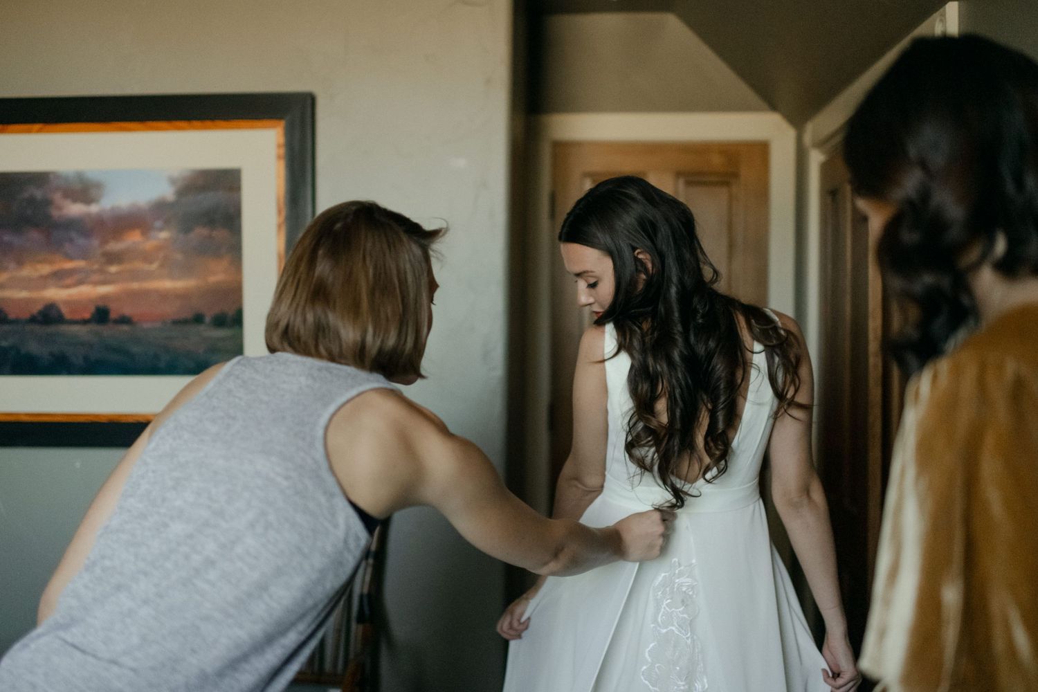 Helping hands assist with wedding dress preparations in front of sunset artwork.