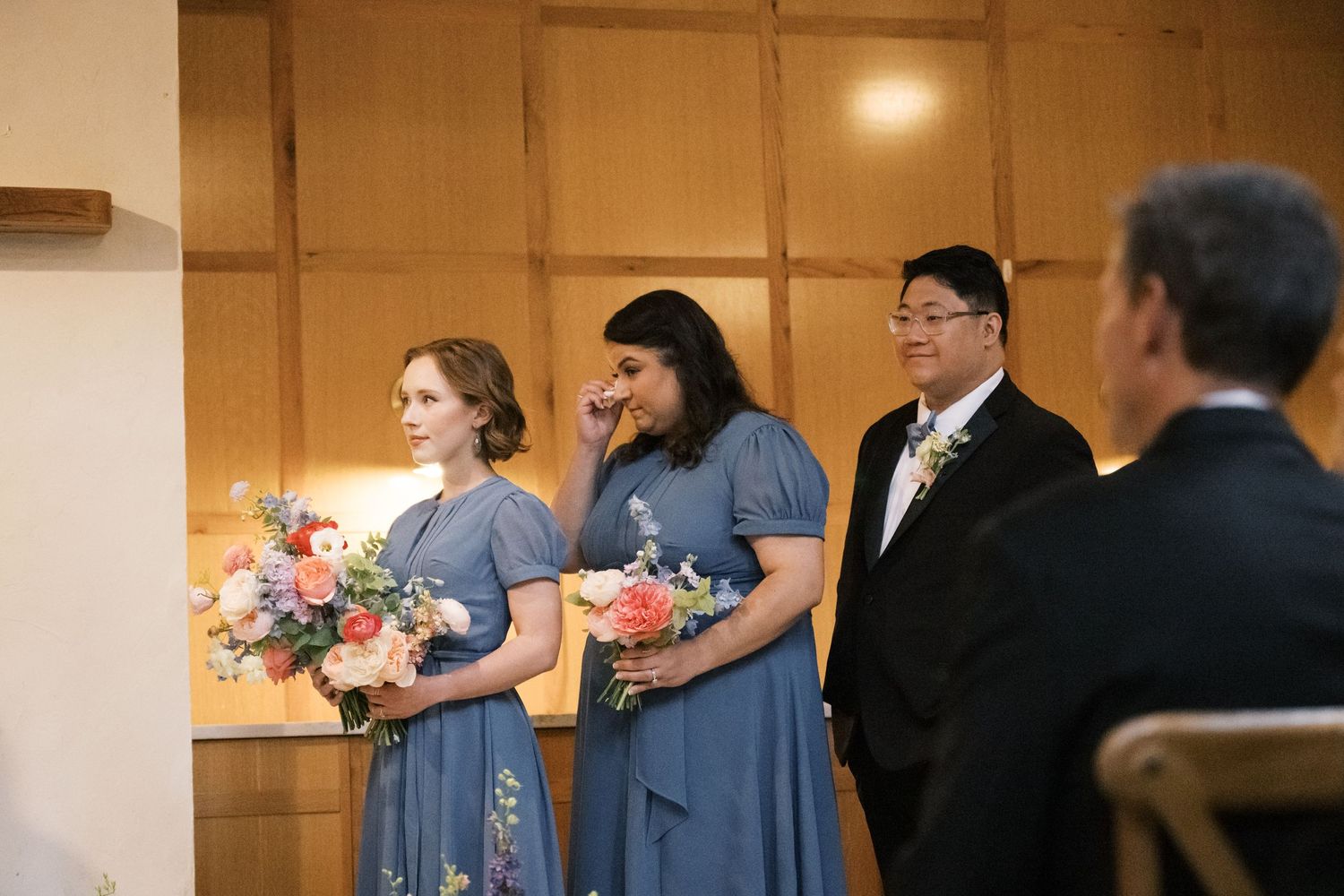 Wedding party members in blue dresses hold colorful bouquets during ceremony.