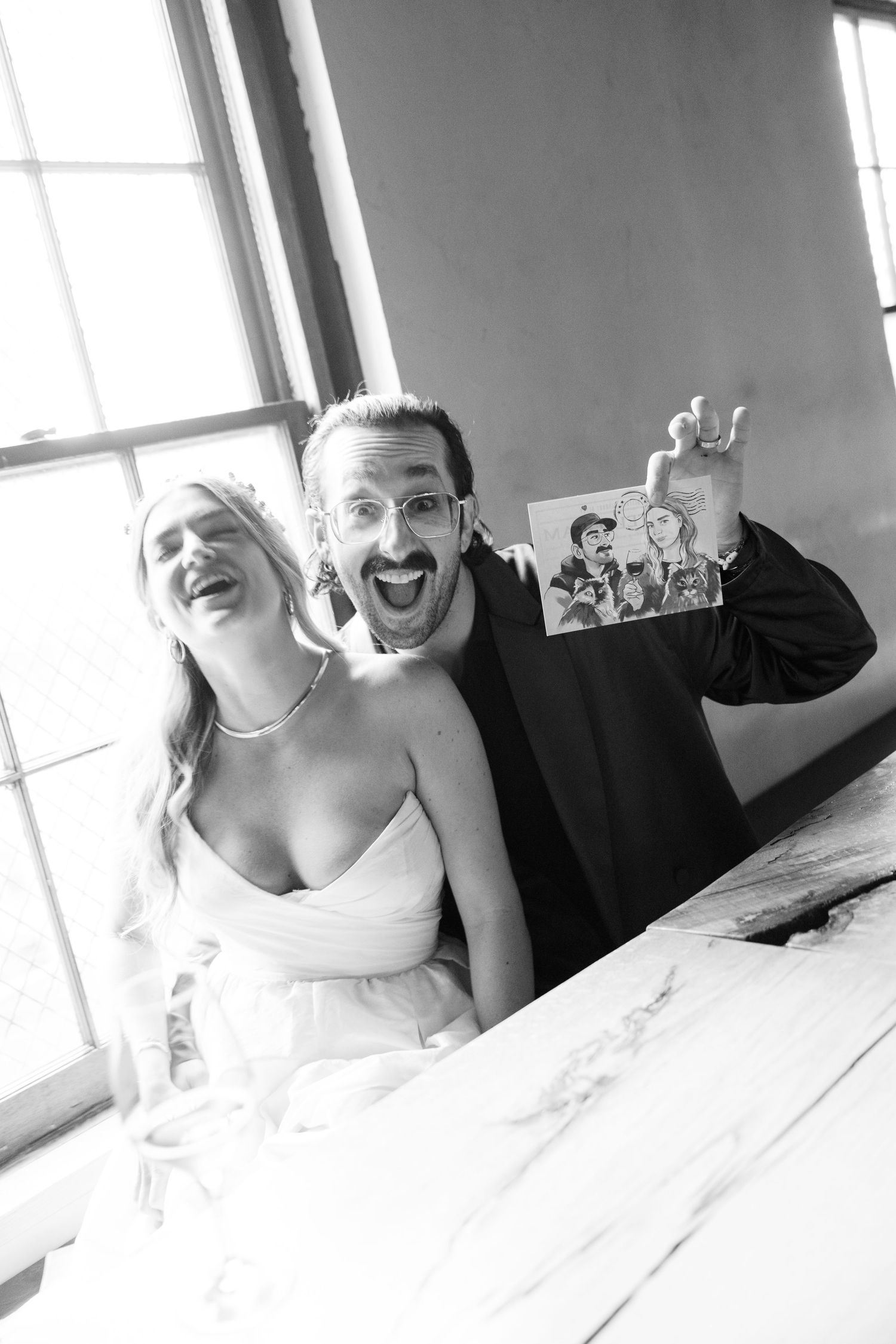 A joyful candid black and white wedding photo showing a couple laughing while holding up an illustration.