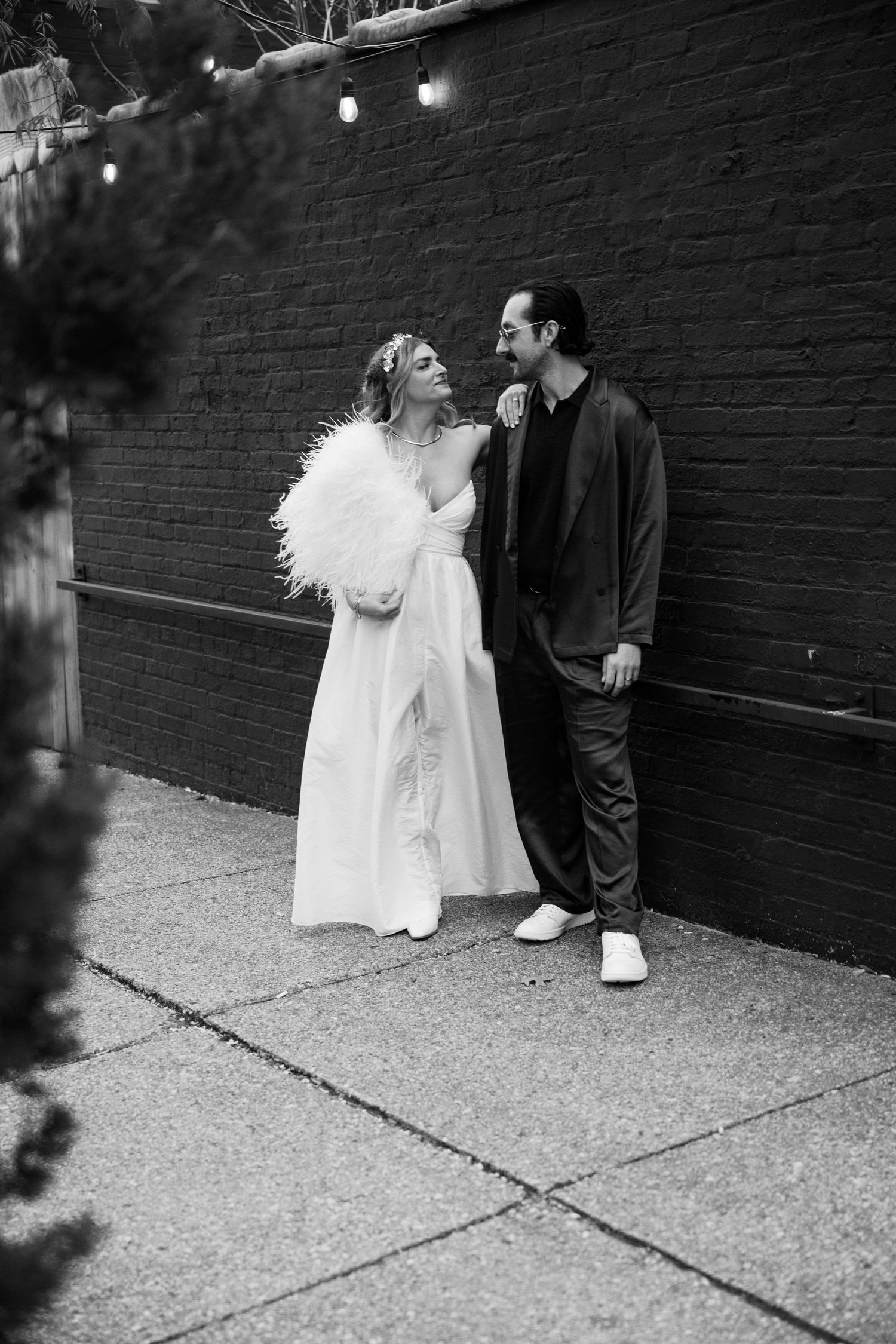 A black and white photo of a couple posing by a brick wall with string lights at Fadensonnen in Baltimore