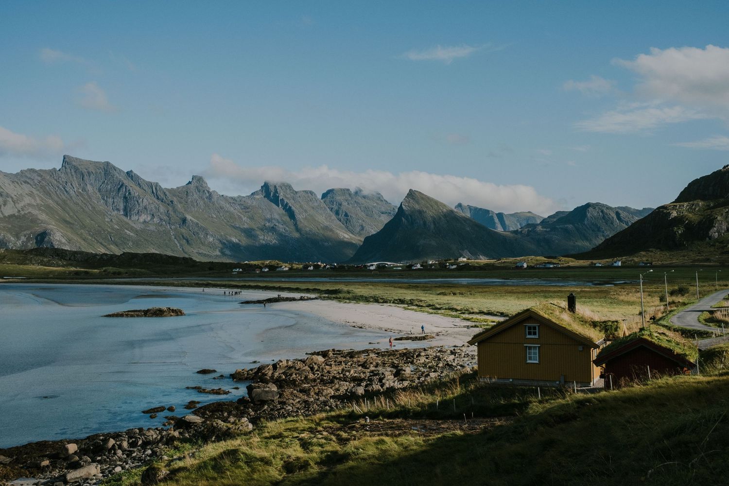 Wooden cabin nestled along fjord shoreline with dramatic mountain range in background.
