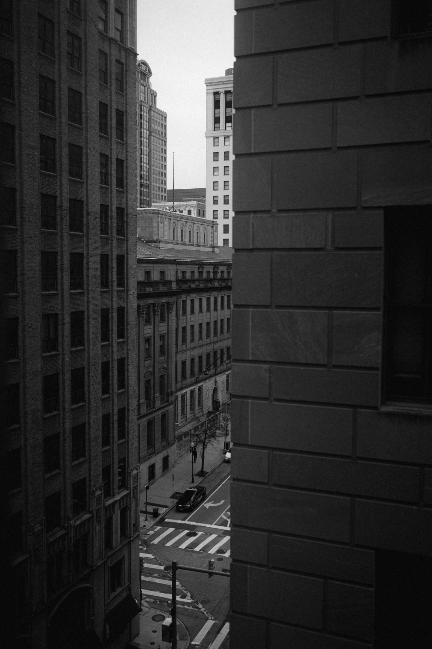 A black and white baltimore city view between tall buildings showing an empty intersection and skyscrapers in the city.