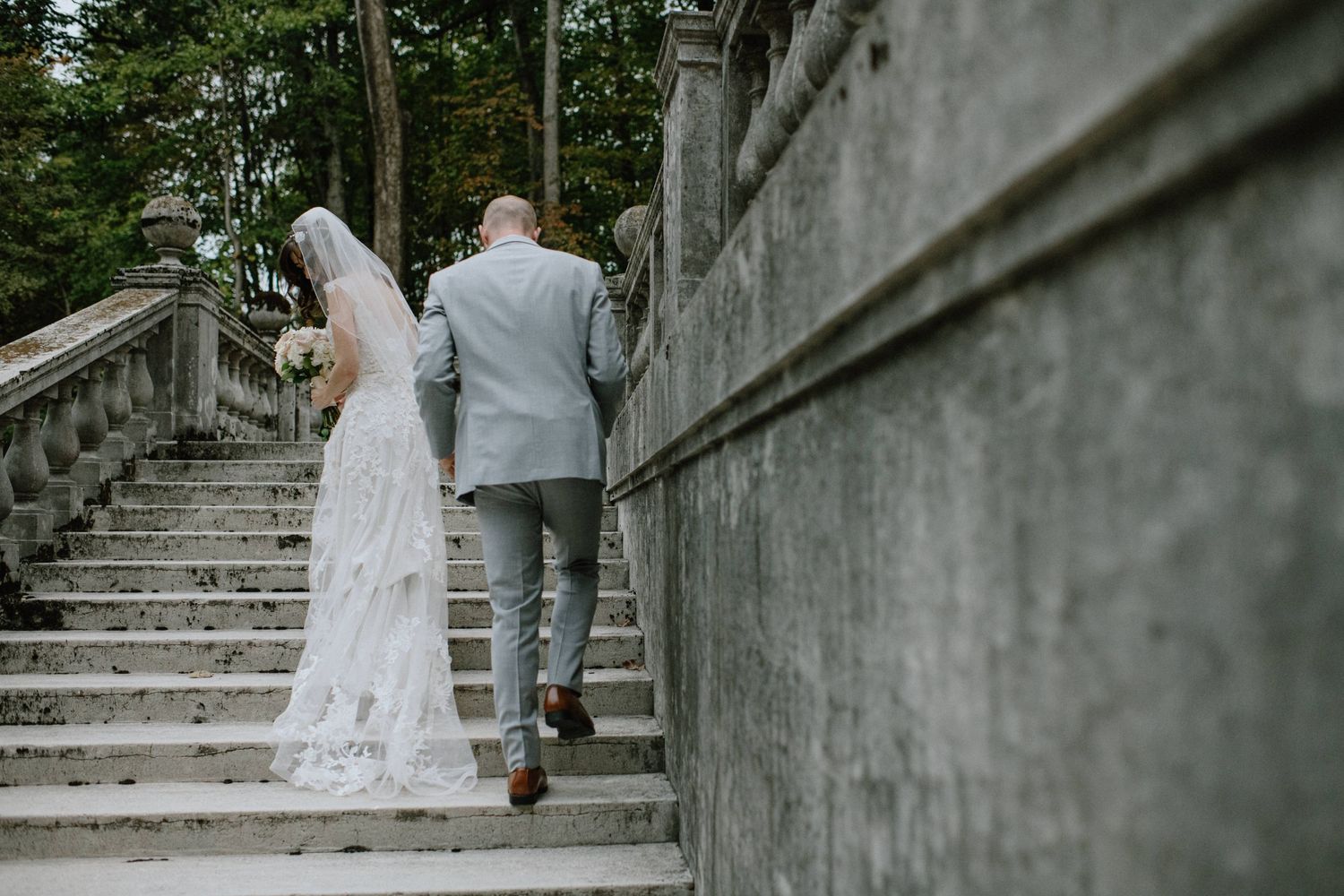Wedding couple climbs elegant stone stairs together surrounded by lush gardens.