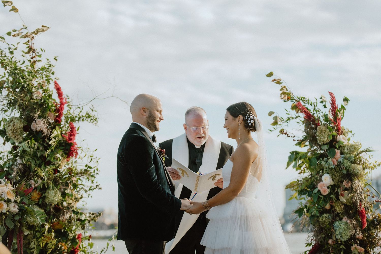 An intimate wedding ceremony moment under an arch adorned with red and white flowers against a cloudy sky.