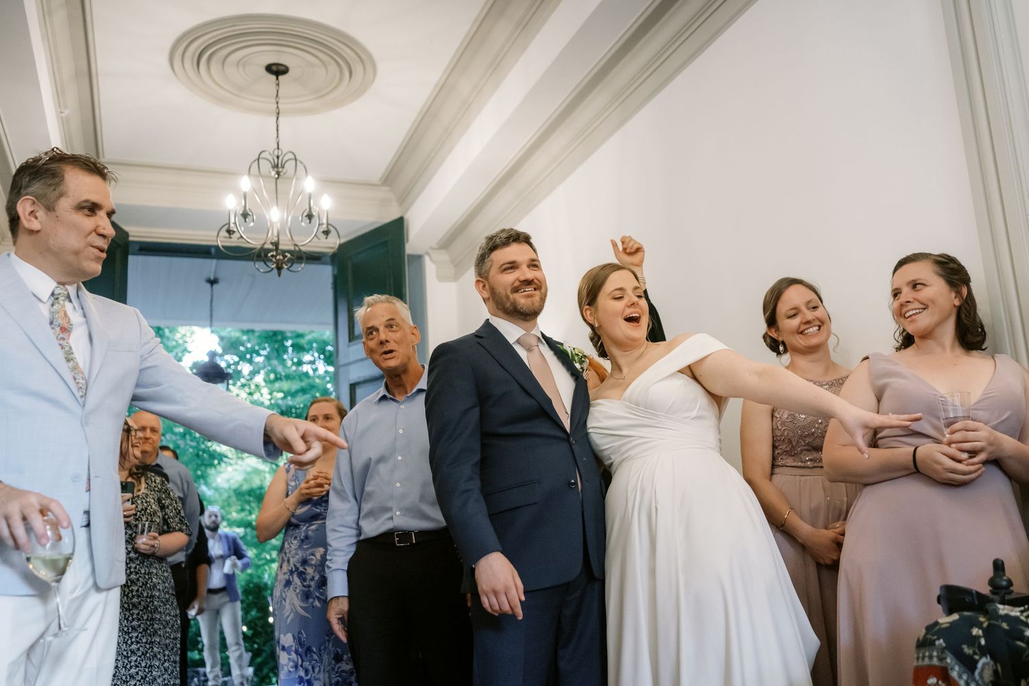 Wedding party celebrating with laughter and drinks in an elegant indoor venue with ornate ceiling details.
