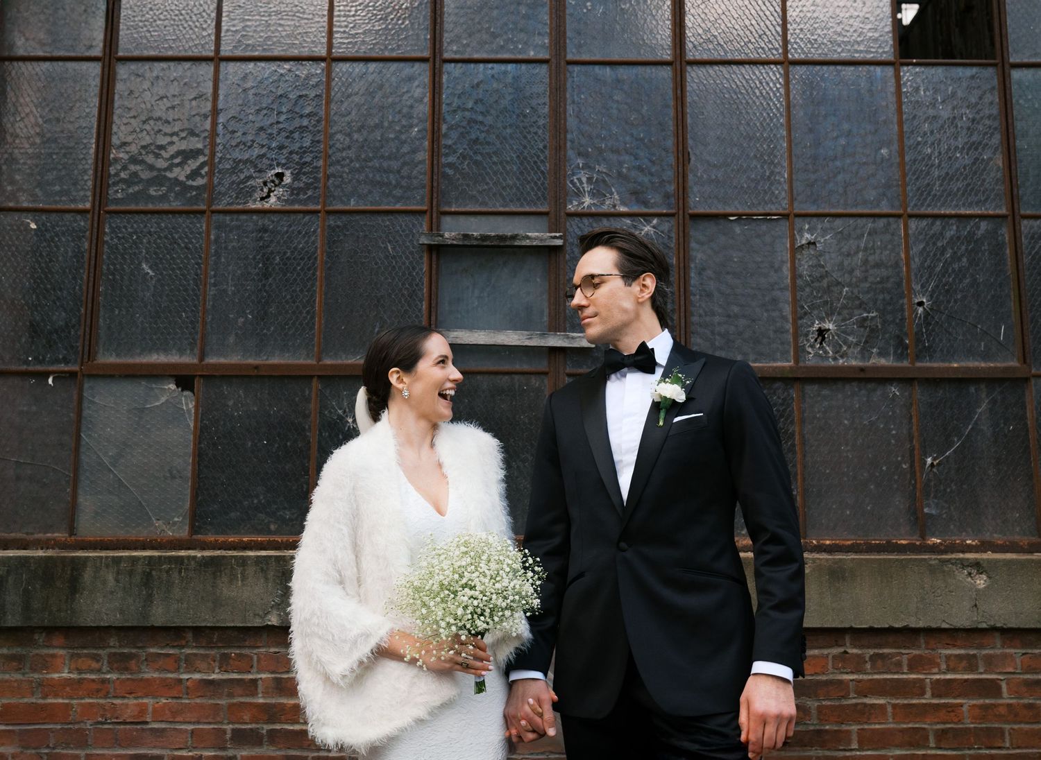 Wedding couple in formal attire standing against industrial window backdrop holding flowers.