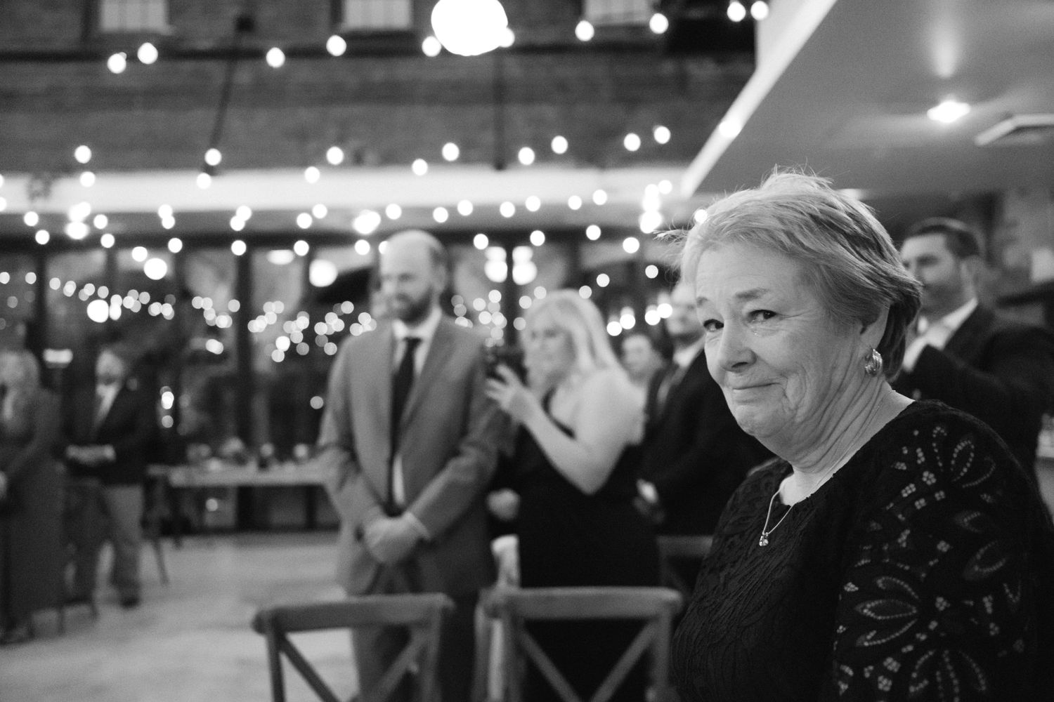 A black and white photo of people at an evening event with twinkling lights in the background.