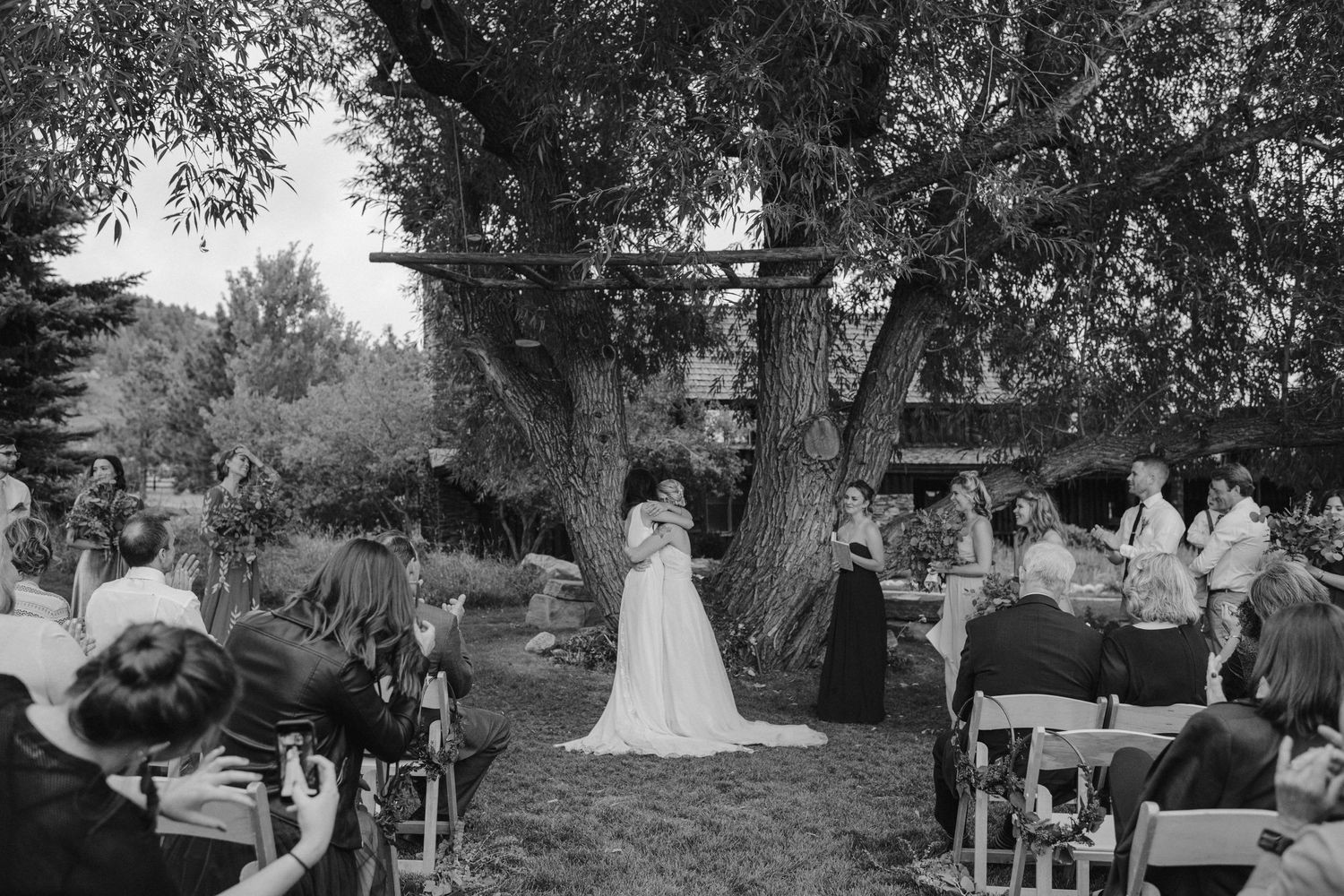 Black and white photo of an outdoor wedding ceremony under a large tree with guests seated in white chairs.