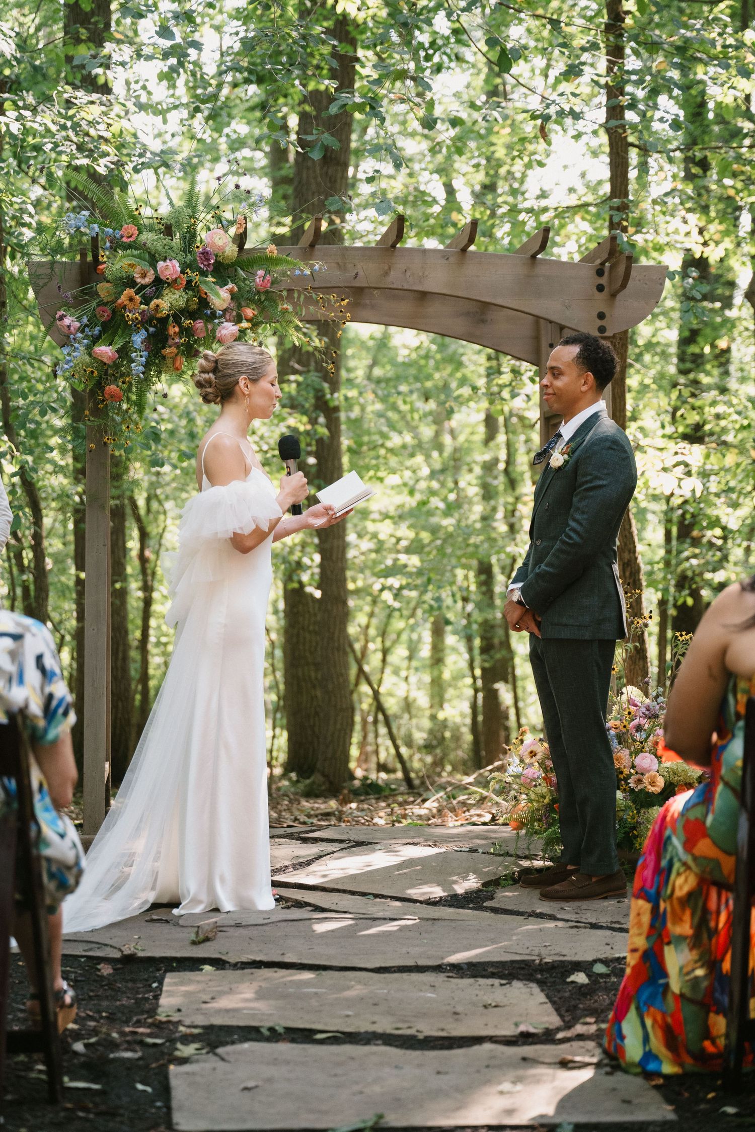 A couple exchanges vows under a rustic wooden arch decorated with colorful flowers in a forest setting.