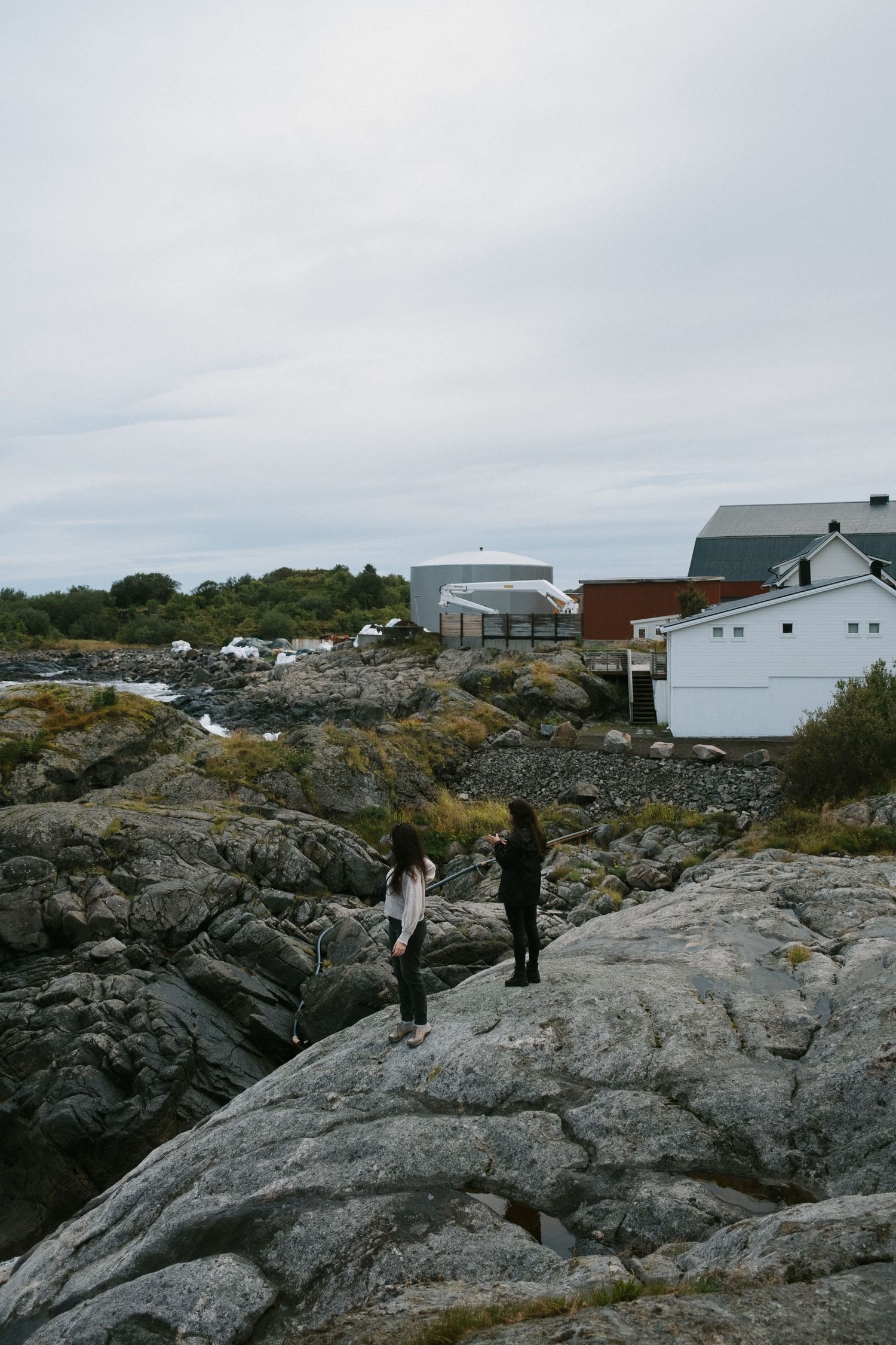 Two people explore rocky coastal terrain with buildings and structures visible in the background on an overcast day.