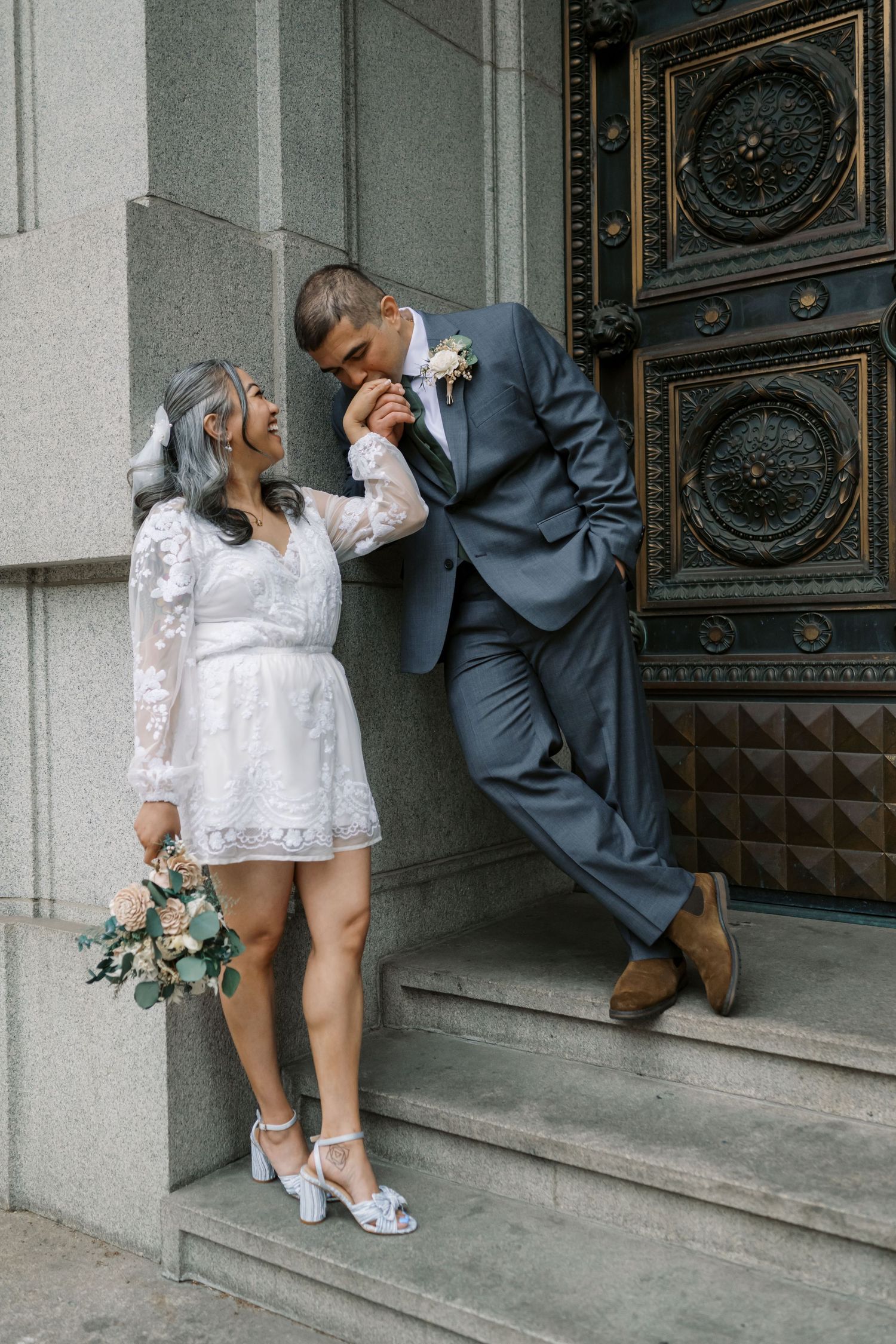 A couple in wedding attire share a romantic moment on stone steps next to ornate vintage doors.