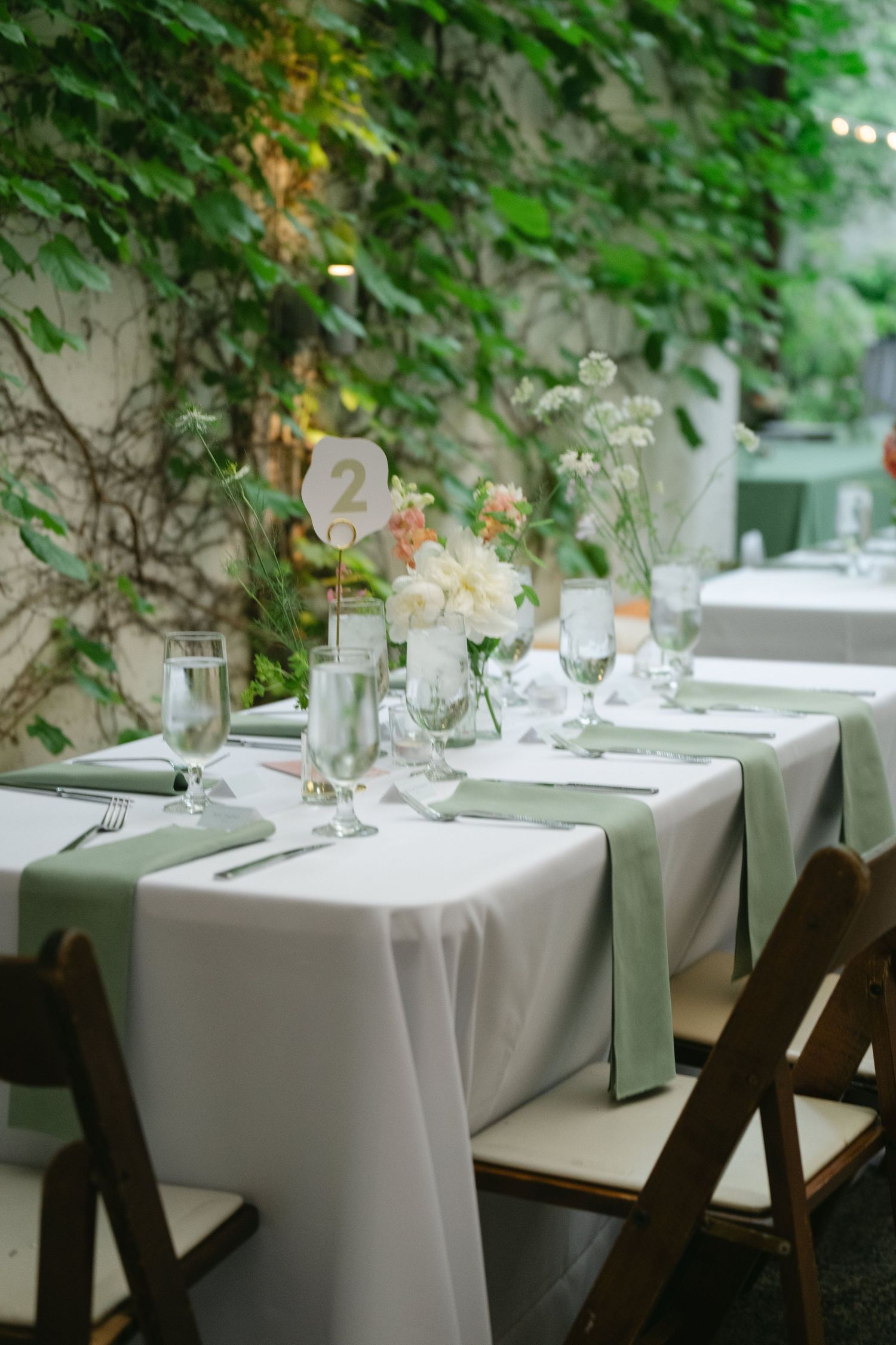 Elegant dining tables with white linens and delicate floral centerpieces set against a backdrop of green foliage.