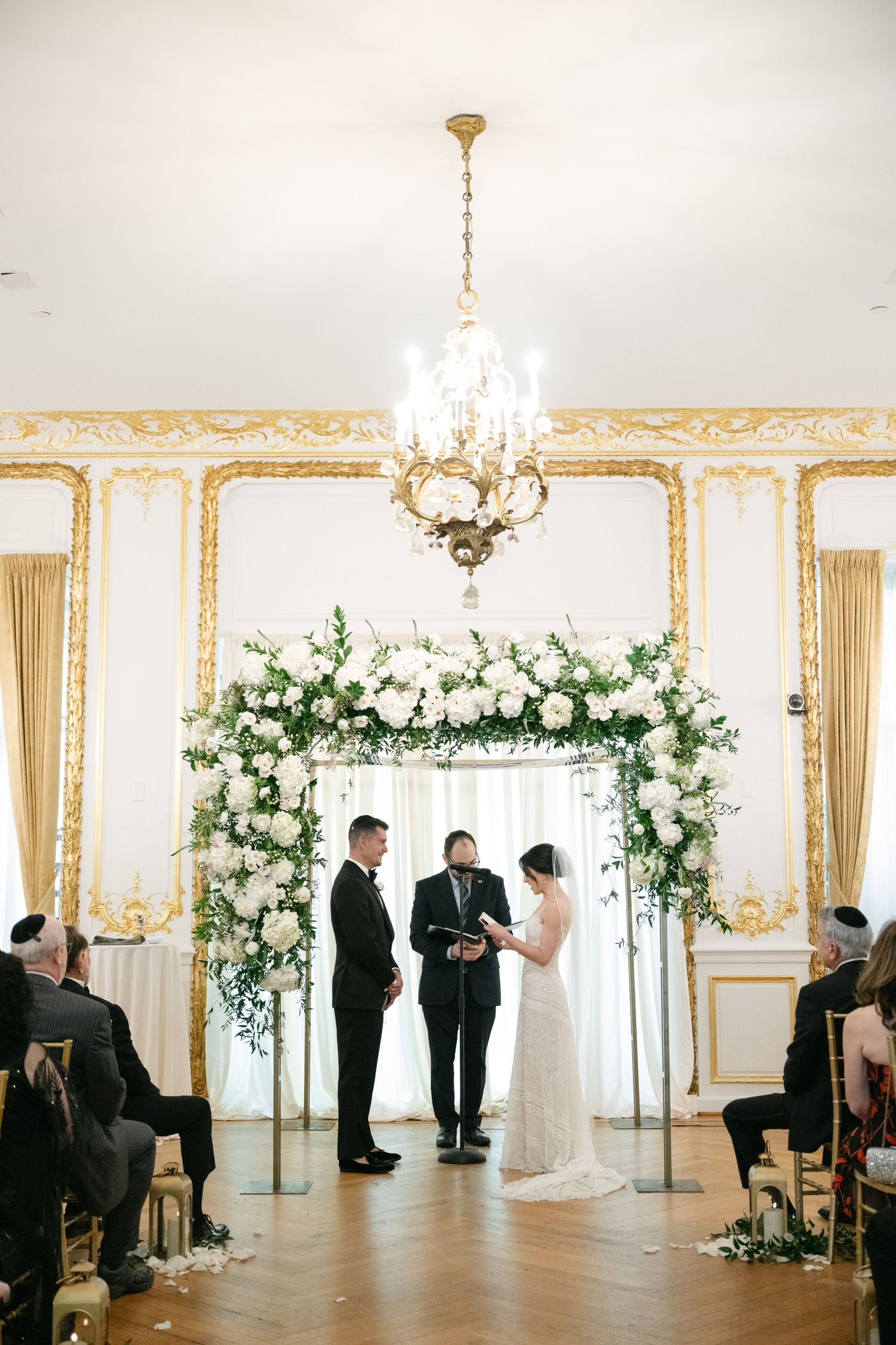 Elegant wedding ceremony setup with white floral arch and crystal chandelier in gilded ballroom.