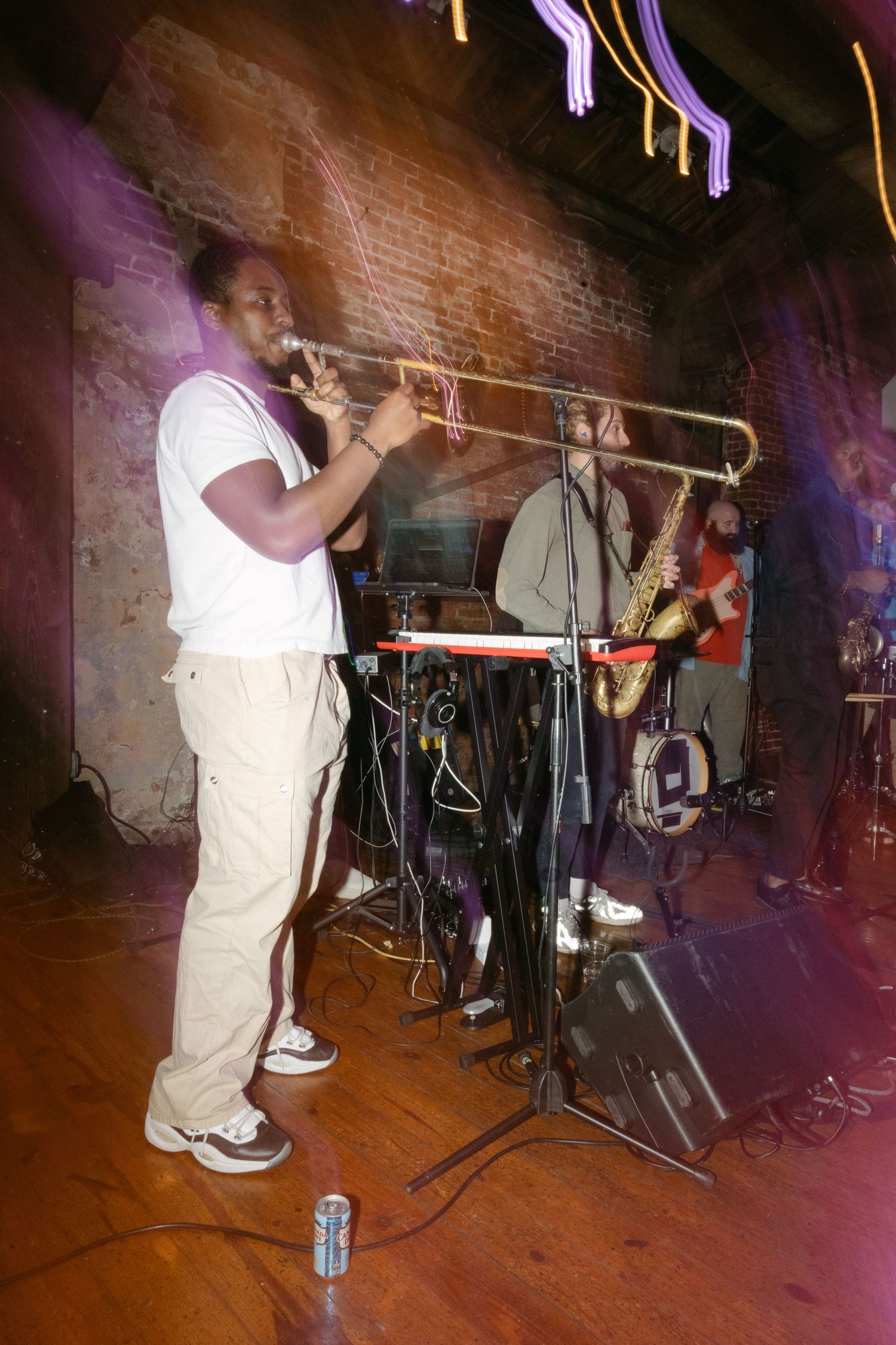 A musician plays trombone on stage in a dimly lit venue with brick walls visible in the background.
