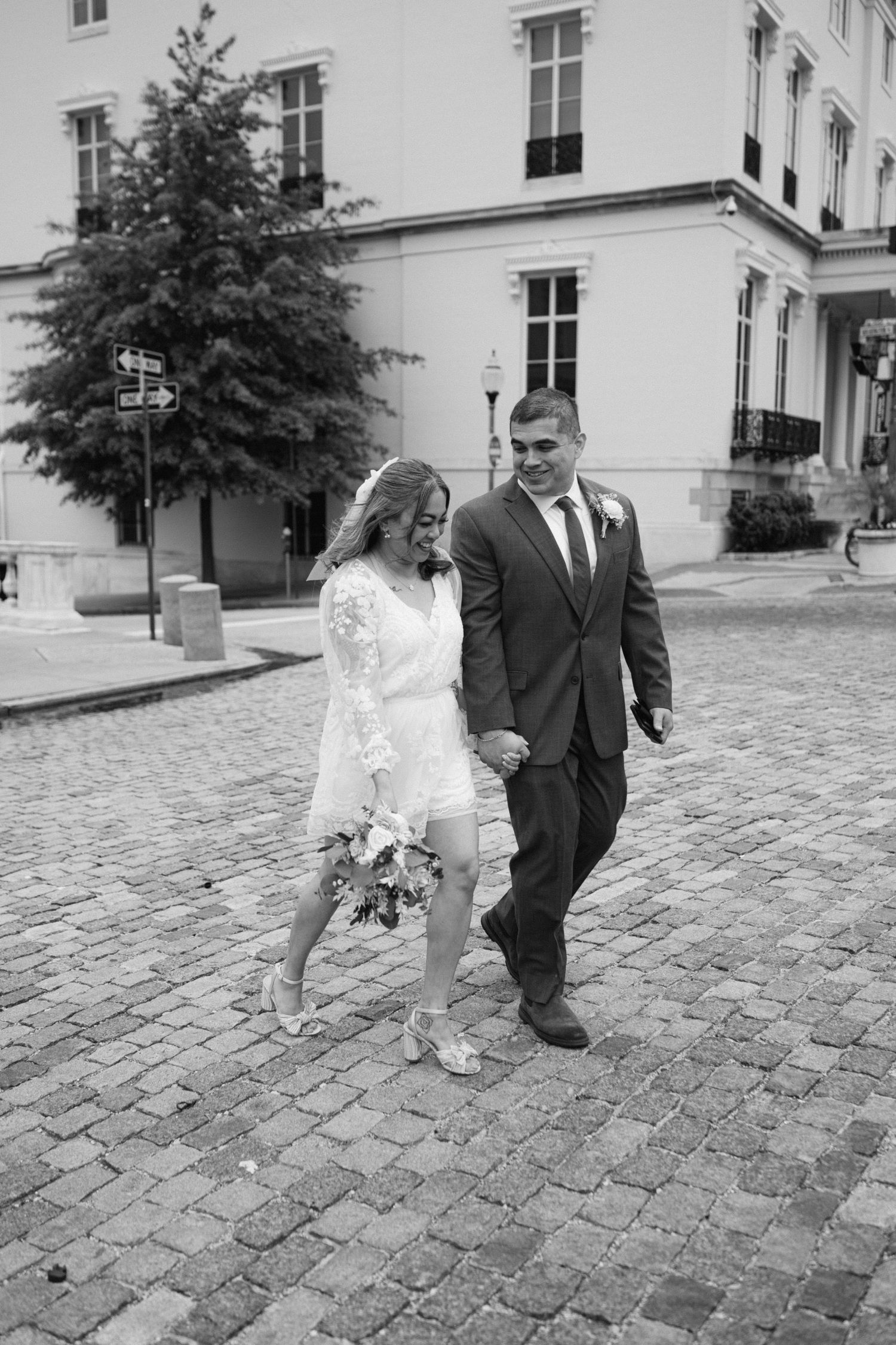 A black and white wedding photo showing a couple walking hand in hand on a cobblestone street near a historic building.